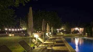 Ein Poolbereich im Freien bei Nacht mit Liegestühlen und Sonnenschirmen, einer Pergola mit Vorhängen sowie einer Steinmauer.
