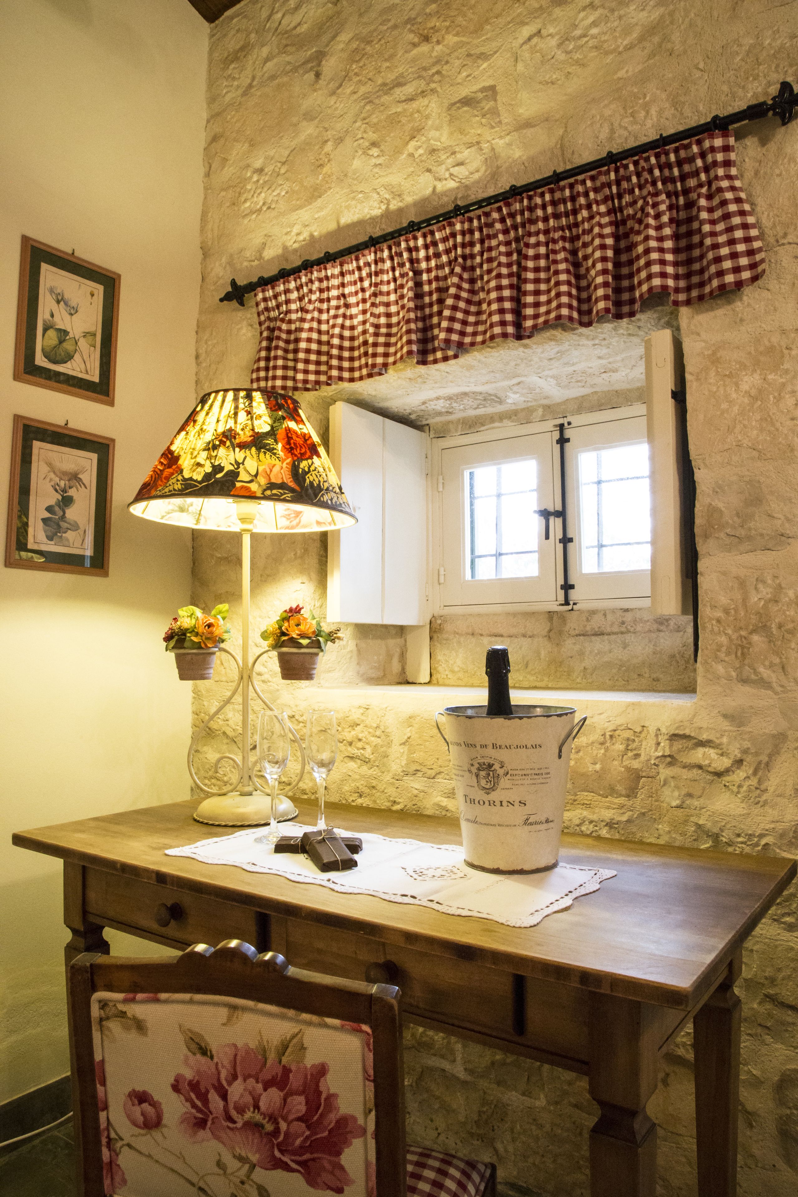 Un'area pranzo rustica con tavolo in legno, lampada floreale, una bottiglia di vino e tenda a quadri.