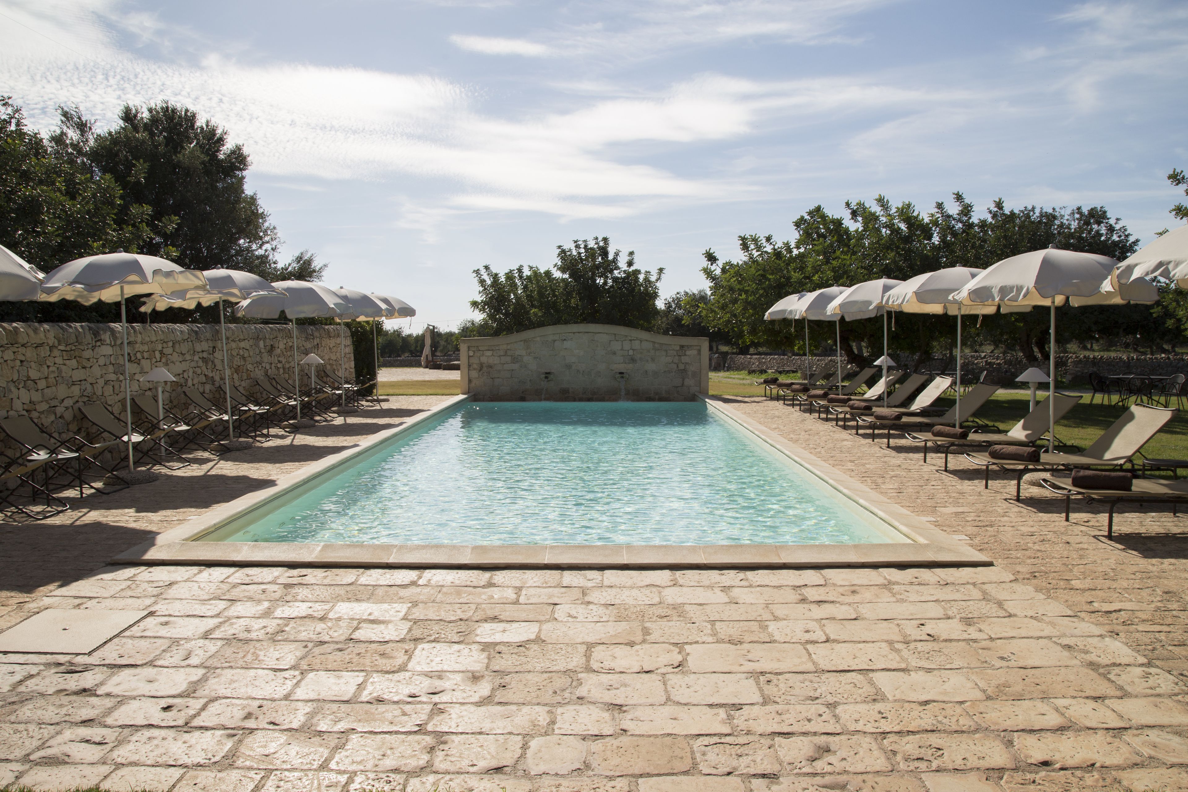 Piscina com espreguiçadeiras e guarda-sóis, cercada por muros de pedra e árvores.
