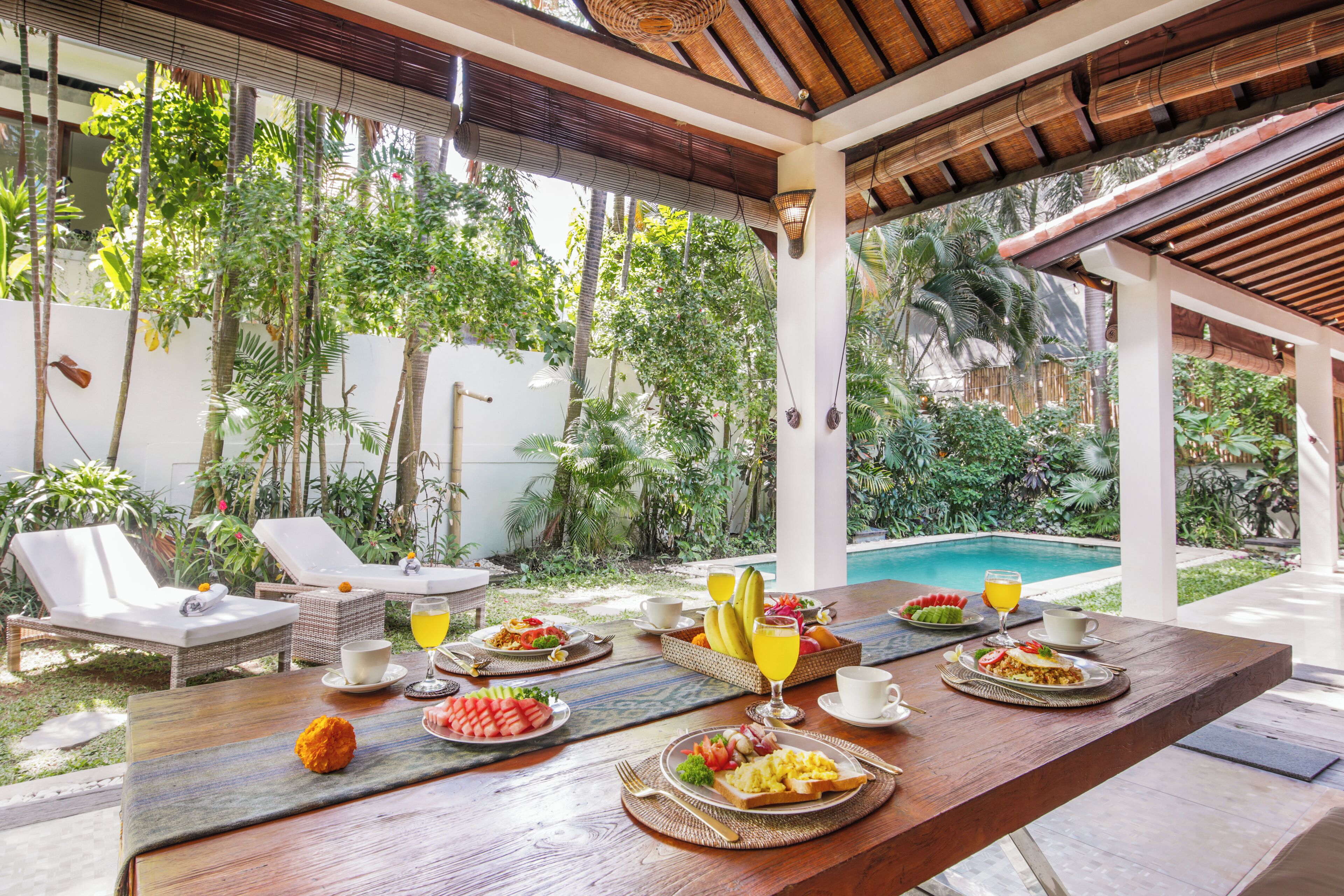 A dining area with a pool view, featuring a wooden table set with breakfast, lounge chairs, and a covered patio.