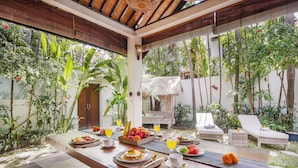 An outdoor dining area with a wooden table set for breakfast, surrounded by lush tropical plants and a thatched-roof structure. - Villa Koala (Seminyak)