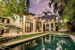 Un área junto a la piscina con un porche cubierto, mobiliario exterior y abundante vegetación.