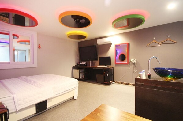 Un dormitorio con cama, escritorio, televisor, espejo, lavamanos y ventana.