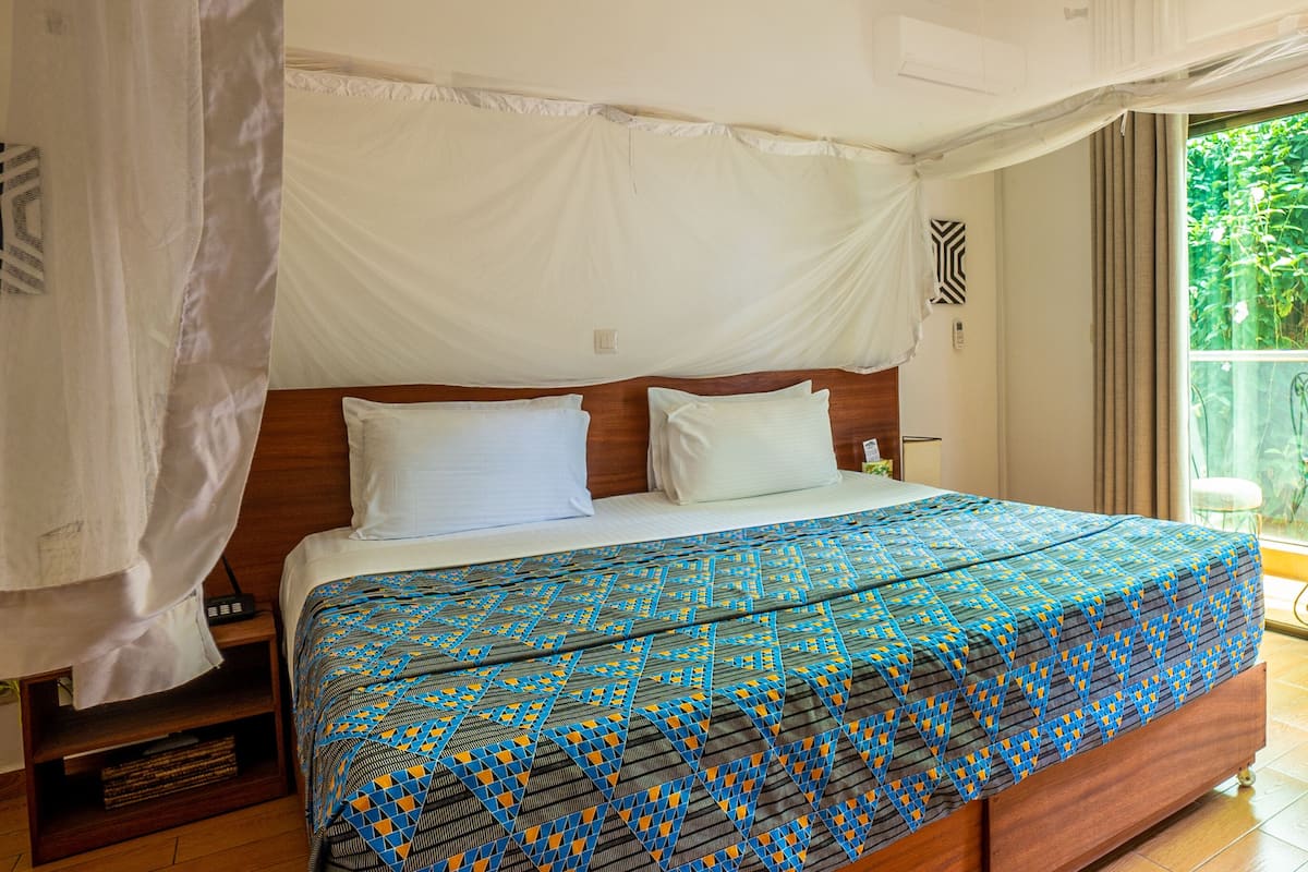 Une chambre avec un grand lit, une tête de lit en bois, une moustiquaire et une vue sur un jardin, accessible par une porte-fenêtre coulissante.