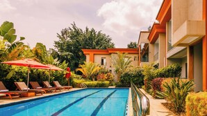 Piscina con sillones de descanso y sombrillas, rodeada de exuberante follaje y un edificio de diseño moderno.