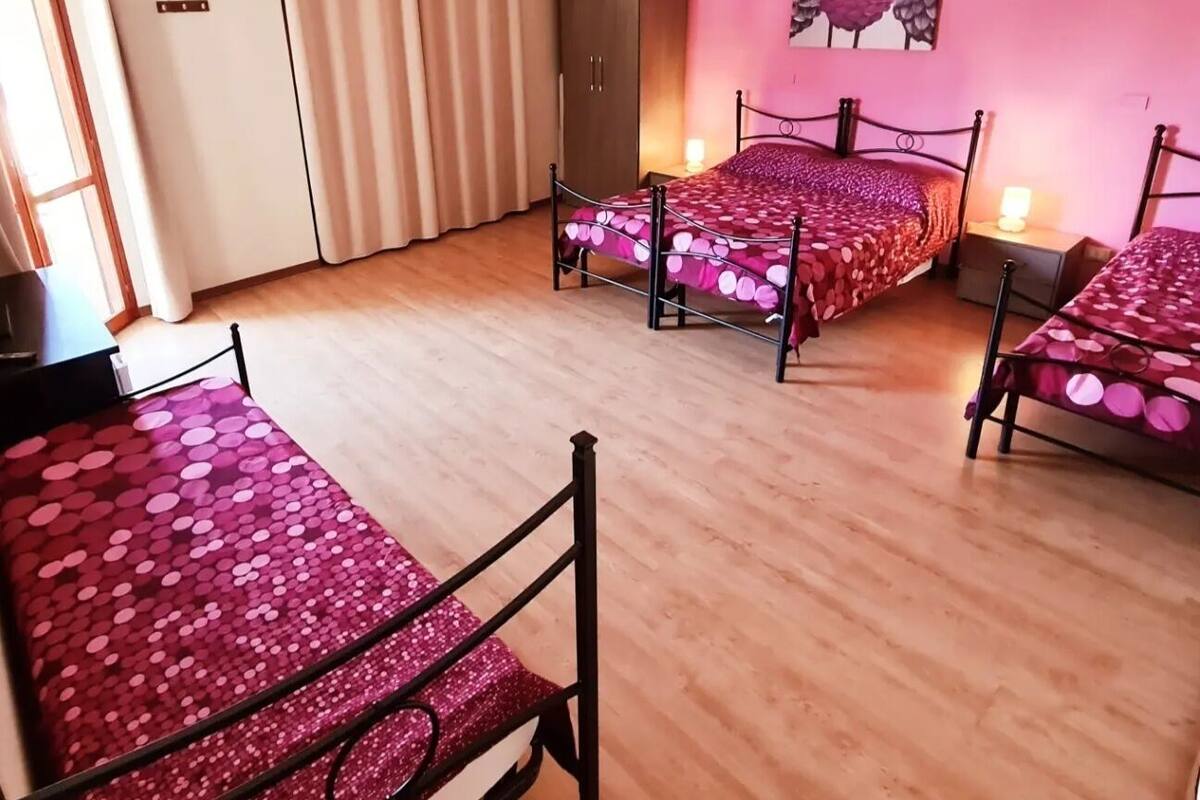 Una habitación con tres camas de estructura metálica y ropa de cama rosa, un armario de madera, una puerta con cortinas y un aire acondicionado instalado en la pared.