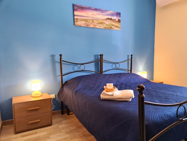 Un dormitorio con pared azul, una cama con colcha azul, una mesita de noche con lámpara y un cuadro de una escena de playa.