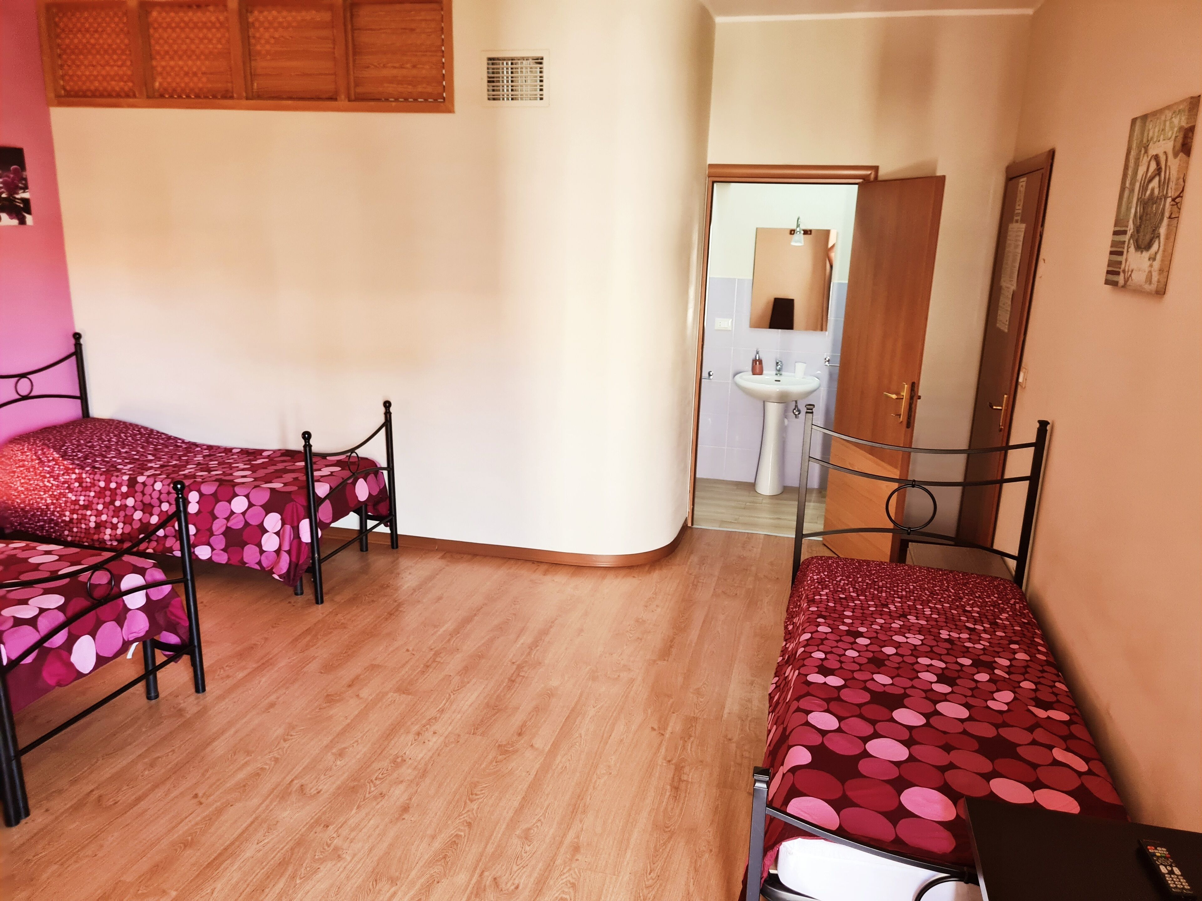 Ein Zimmer mit zwei Einzelbetten, Metallgestellen und roter Bettwäsche, einem Holzboden, einem weißen Waschbecken und einer Tür zu einem weiteren Raum.