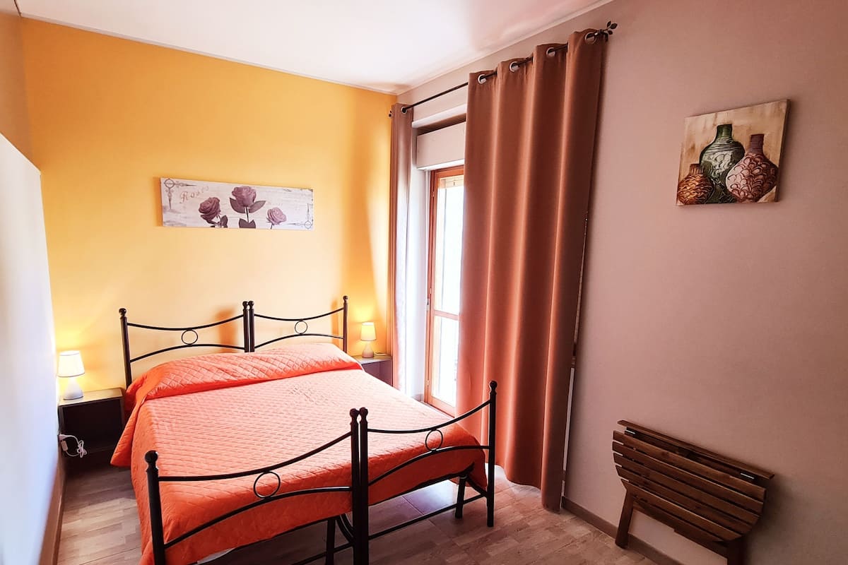 Un dormitorio con cama, una mesita de noche, lámpara, ventana con cortinas y un cuadro en la pared.