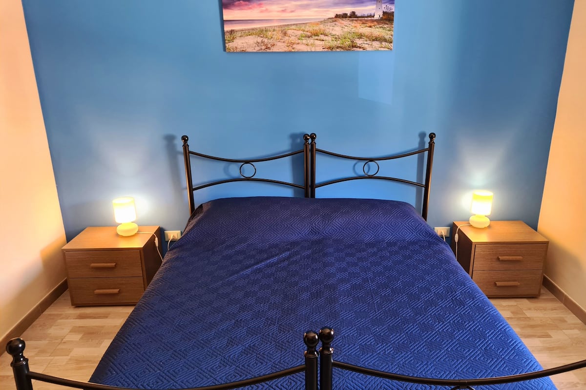 Una cama con colcha azul, dos mesitas de noche con lámparas, un cabecero de madera y un cuadro colgado en la pared.