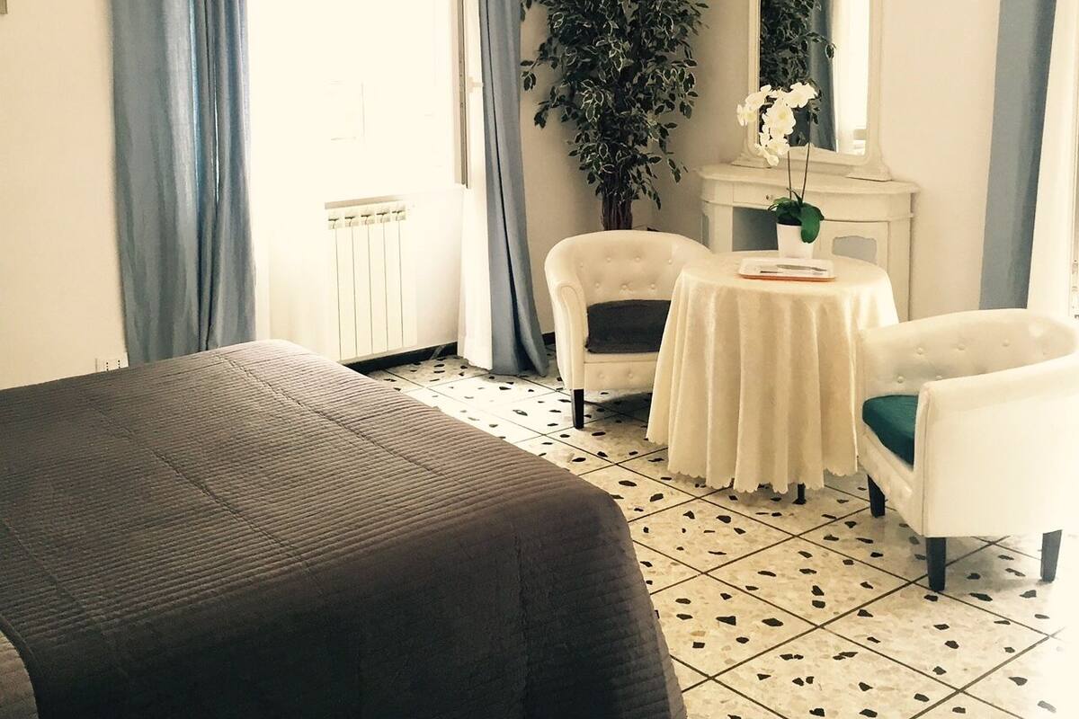 A hotel room with a bed, two white armchairs, a small table with a plant, a mirror, and a chandelier.