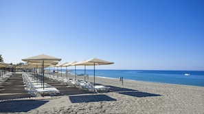 Une plage bordée de rangées de chaises longues blanches et de parasols beiges, un ciel bleu azur et une mer calme.