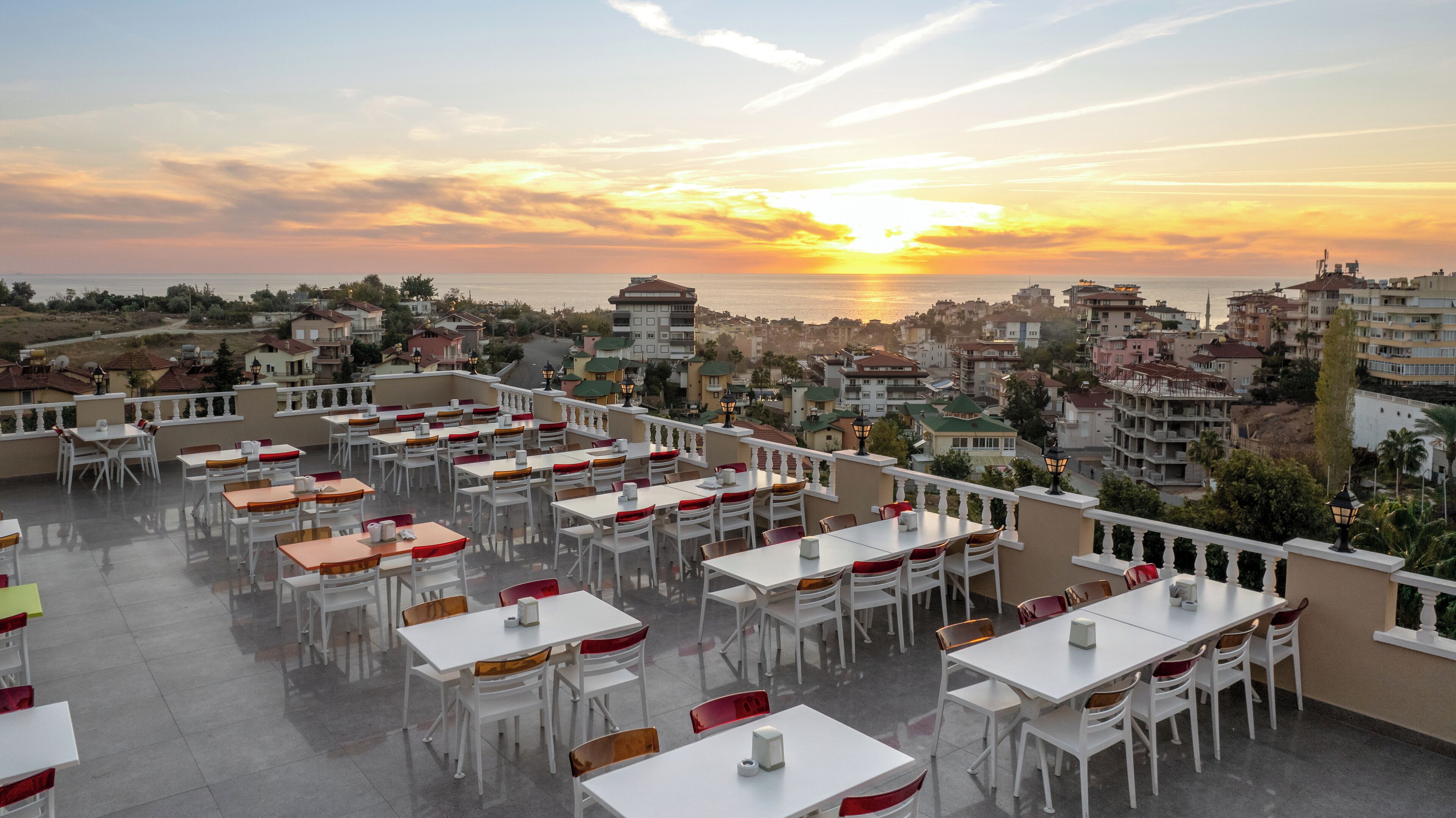 Terraza en la azotea con mesas y sillas, con vistas a una ciudad costera al atardecer.