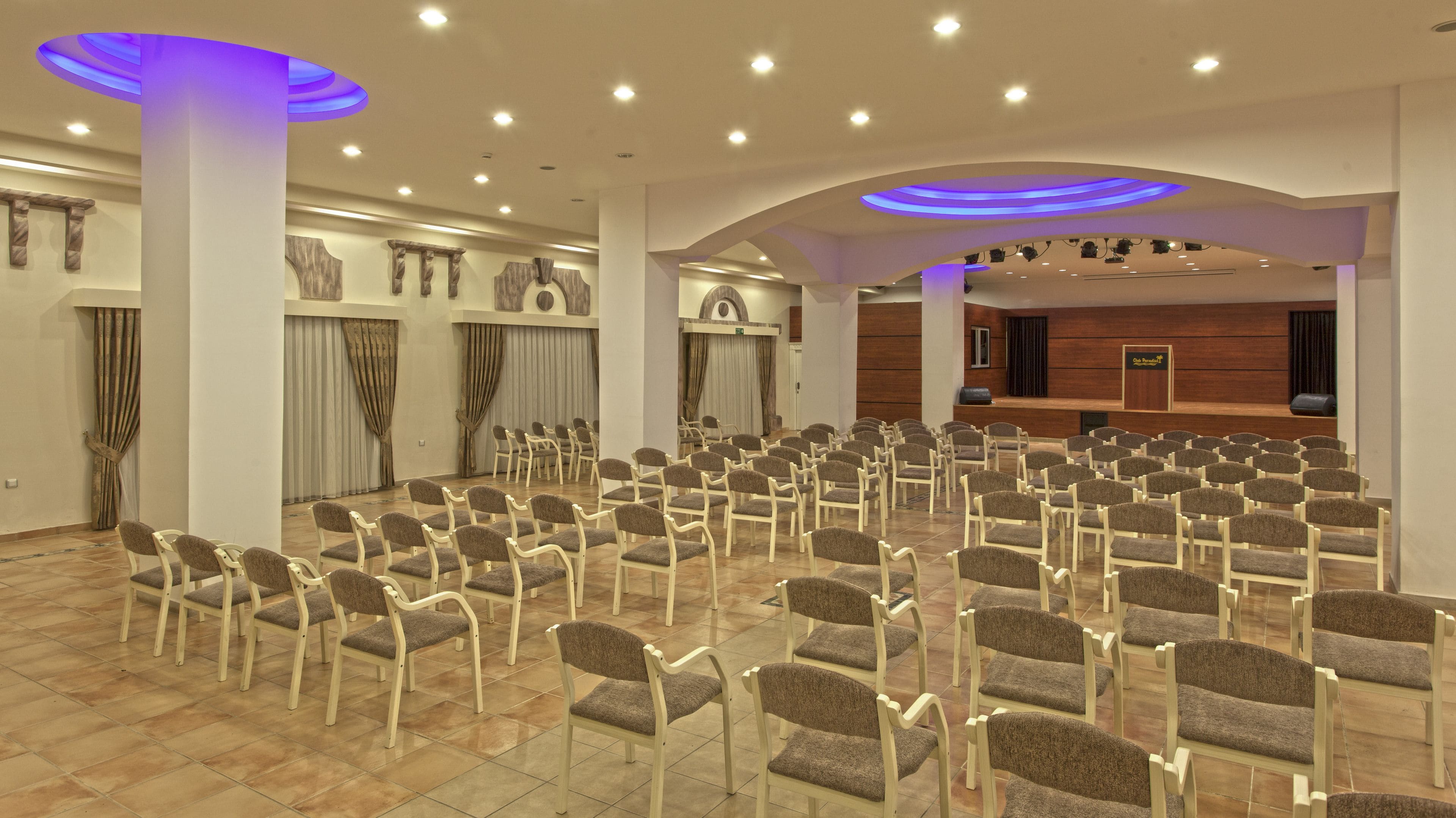A spacious conference room with rows of chairs, a stage, and modern lighting fixtures.