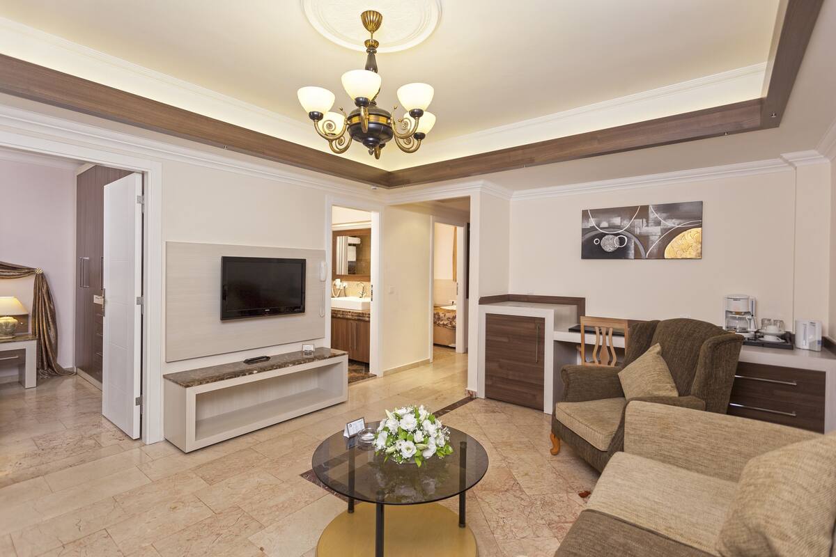 A modern living room with a flat-screen TV, a glass coffee table with flowers, a sofa, and a dining area with a table and chairs.