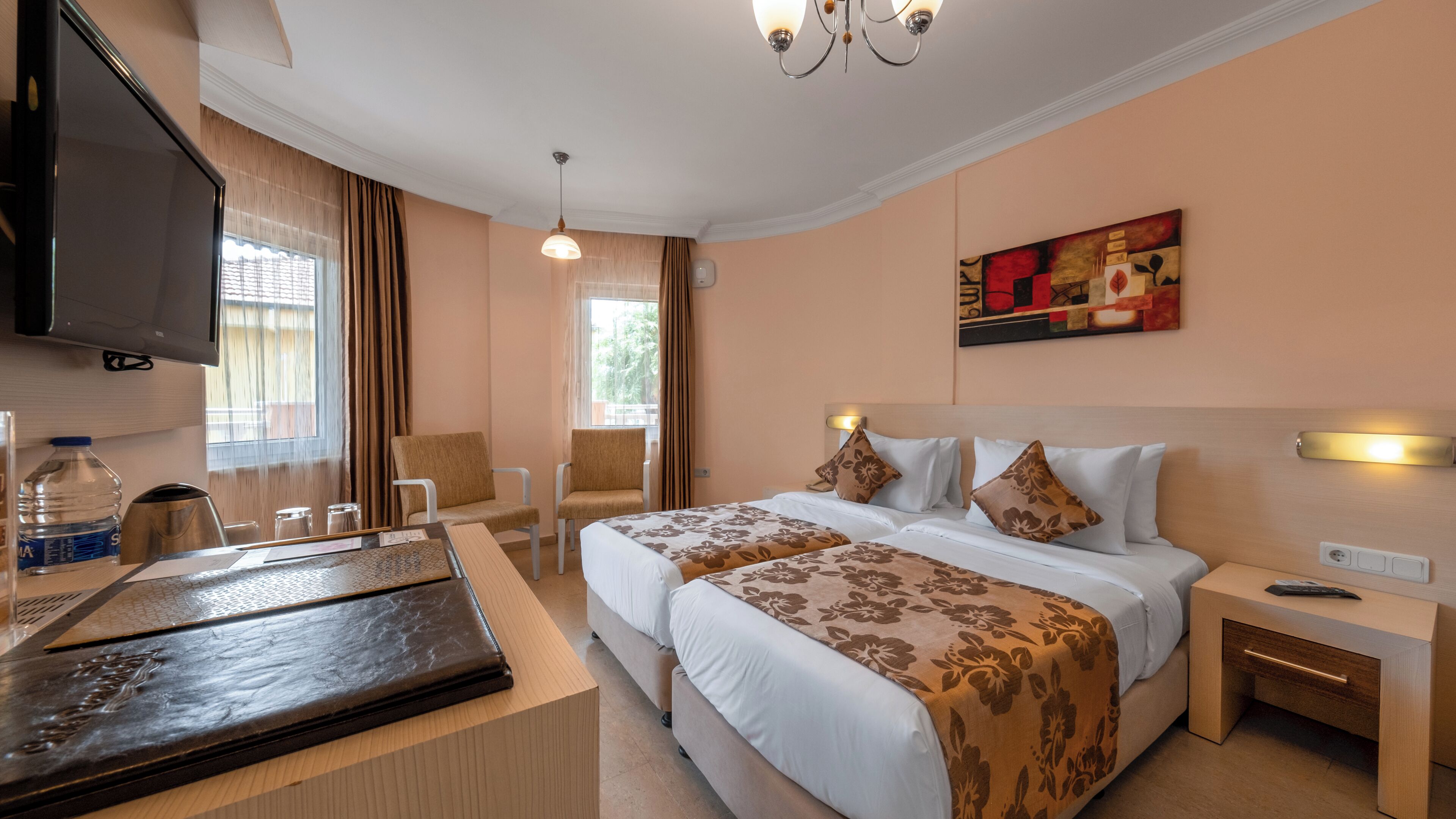 Een hotelkamer met een bed, nachtkastjes, een televisie, een bureau met een fles water, en een schilderij aan de muur.