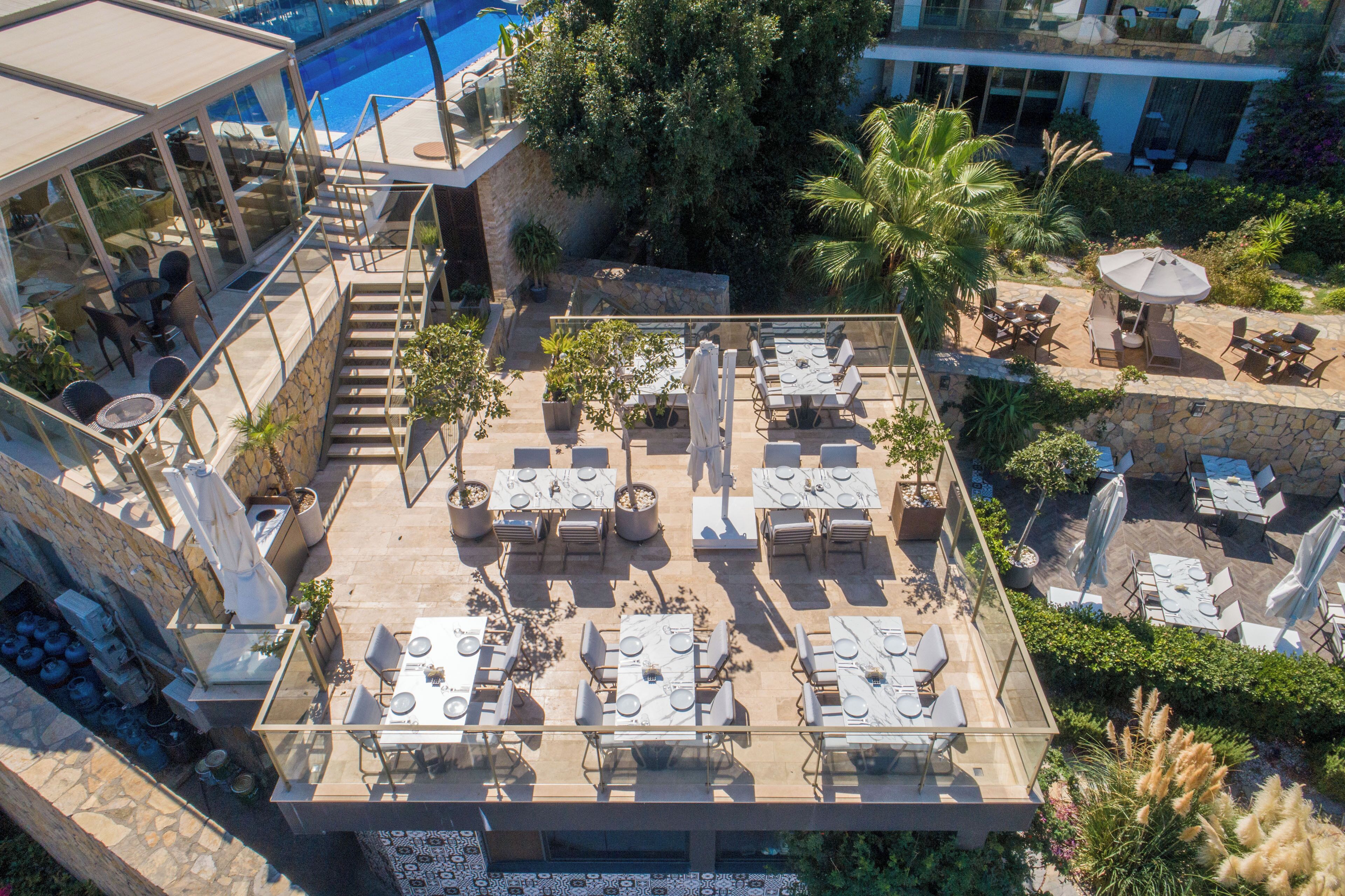 An outdoor dining area with multiple tables and chairs, surrounded by greenery and a pool.