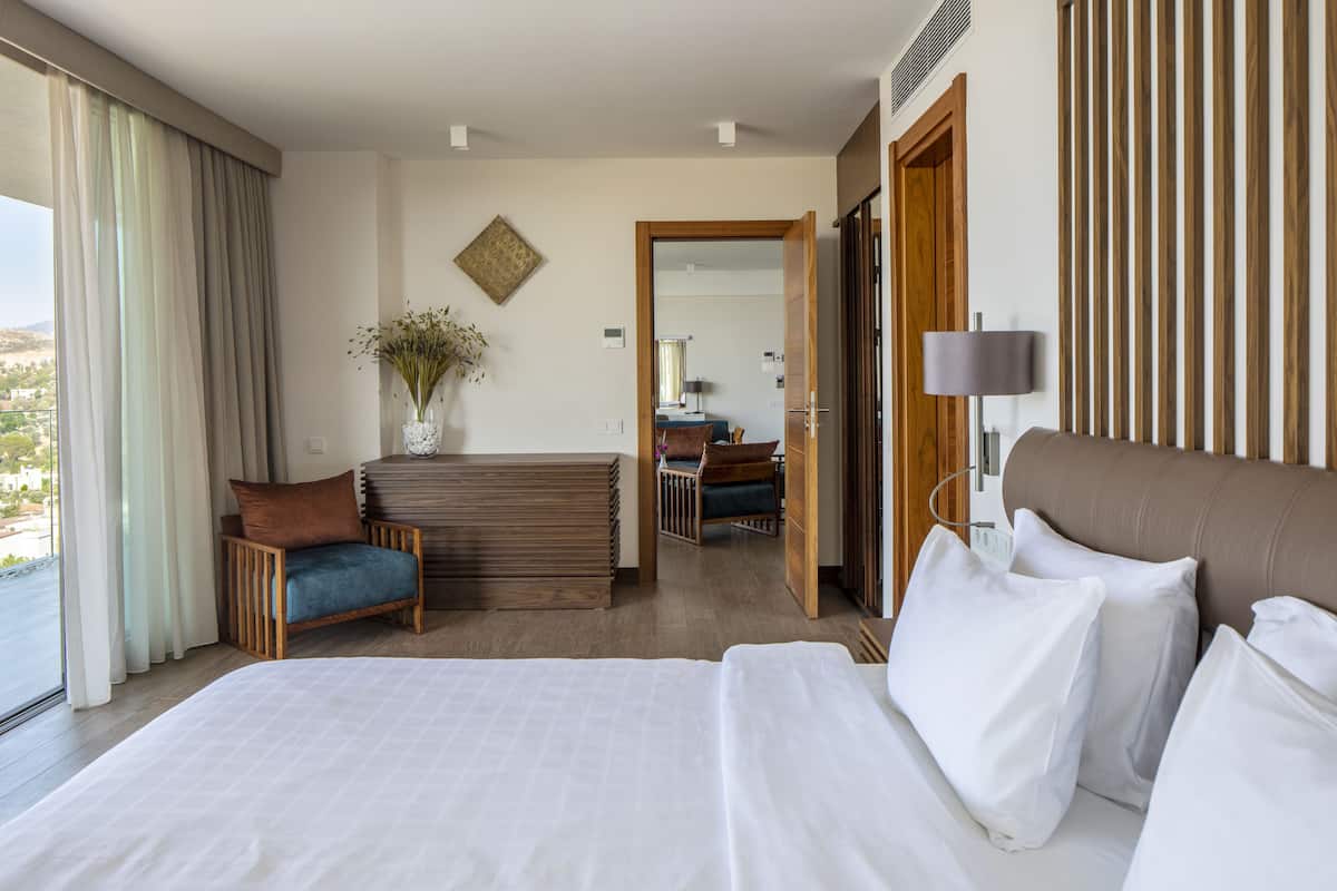 A hotel room with a large bed, a sitting area with a chair and a small table, a view of the outdoors, and a wooden door leading to another room.
