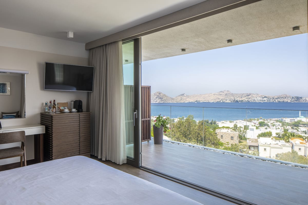 A modern hotel room with a large bed, a desk with a flat-screen TV, a balcony with a view of the sea and cityscape.