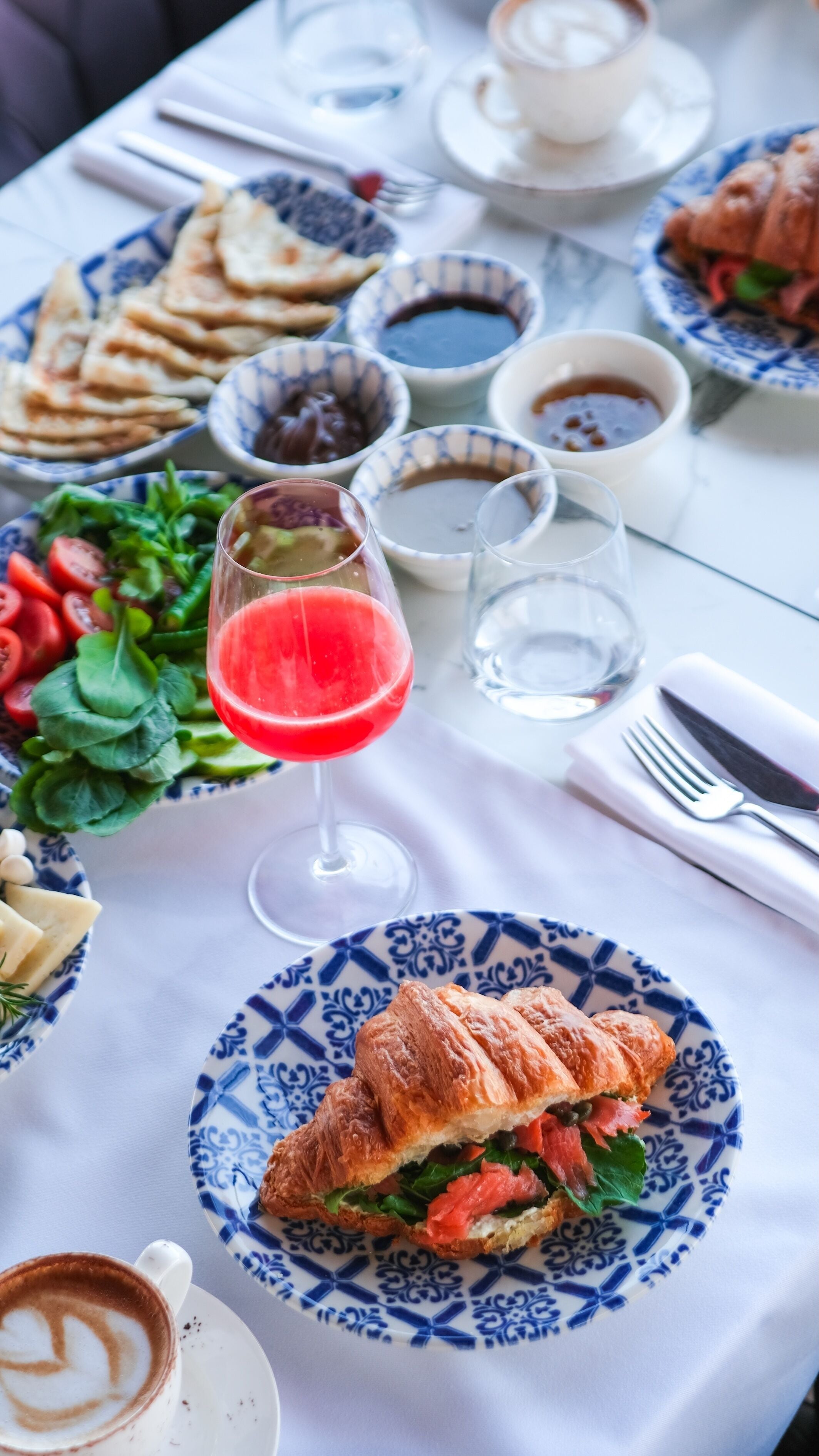Mesa posta com diversos pratos, incluindo um croissant, salada e um copo de bebida vermelha.