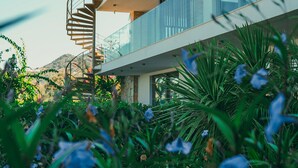 Un edificio moderno con una escalera de caracol, balcones de cristal y abundante vegetación circundante.