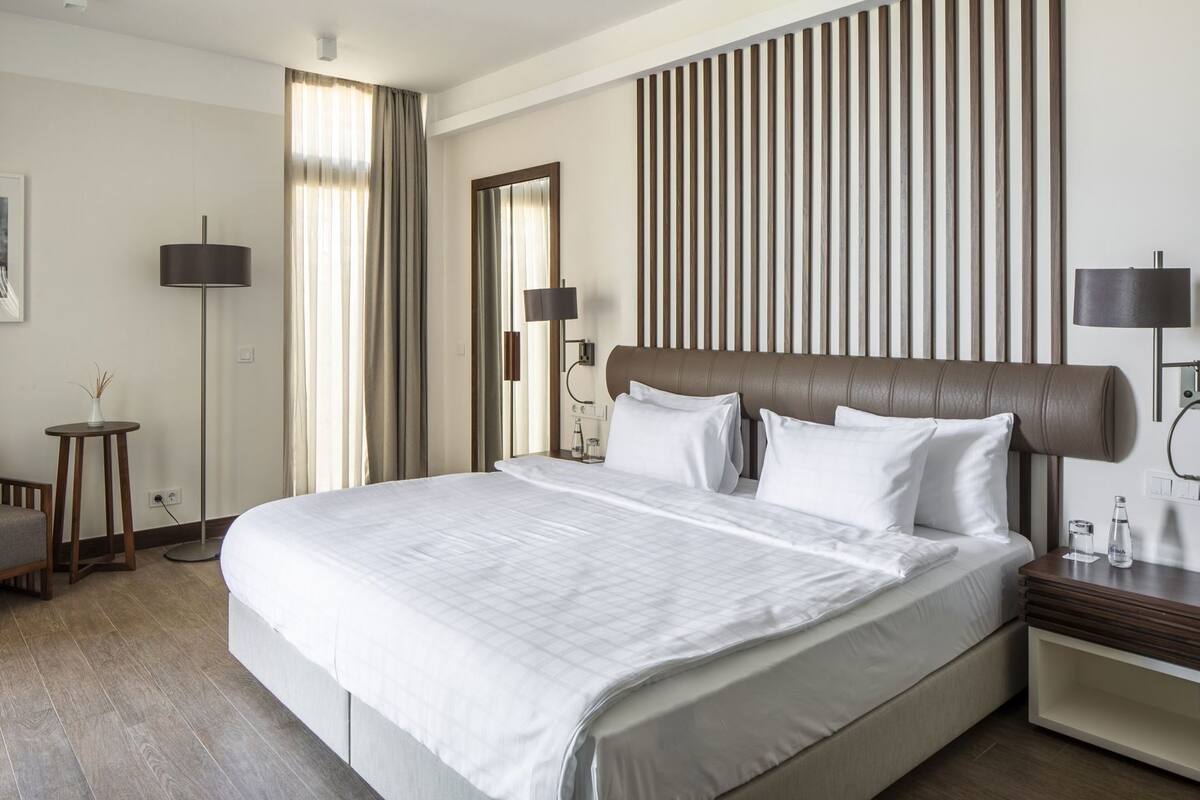 A hotel room with a large bed, bedside tables, a floor lamp, a mirror, and a view of the outside.