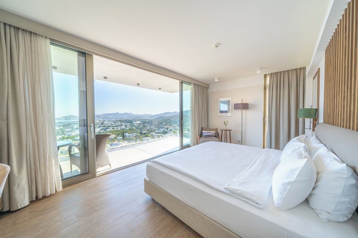 A modern hotel room with a large bed, a sliding glass door leading to a balcony, and a city view.