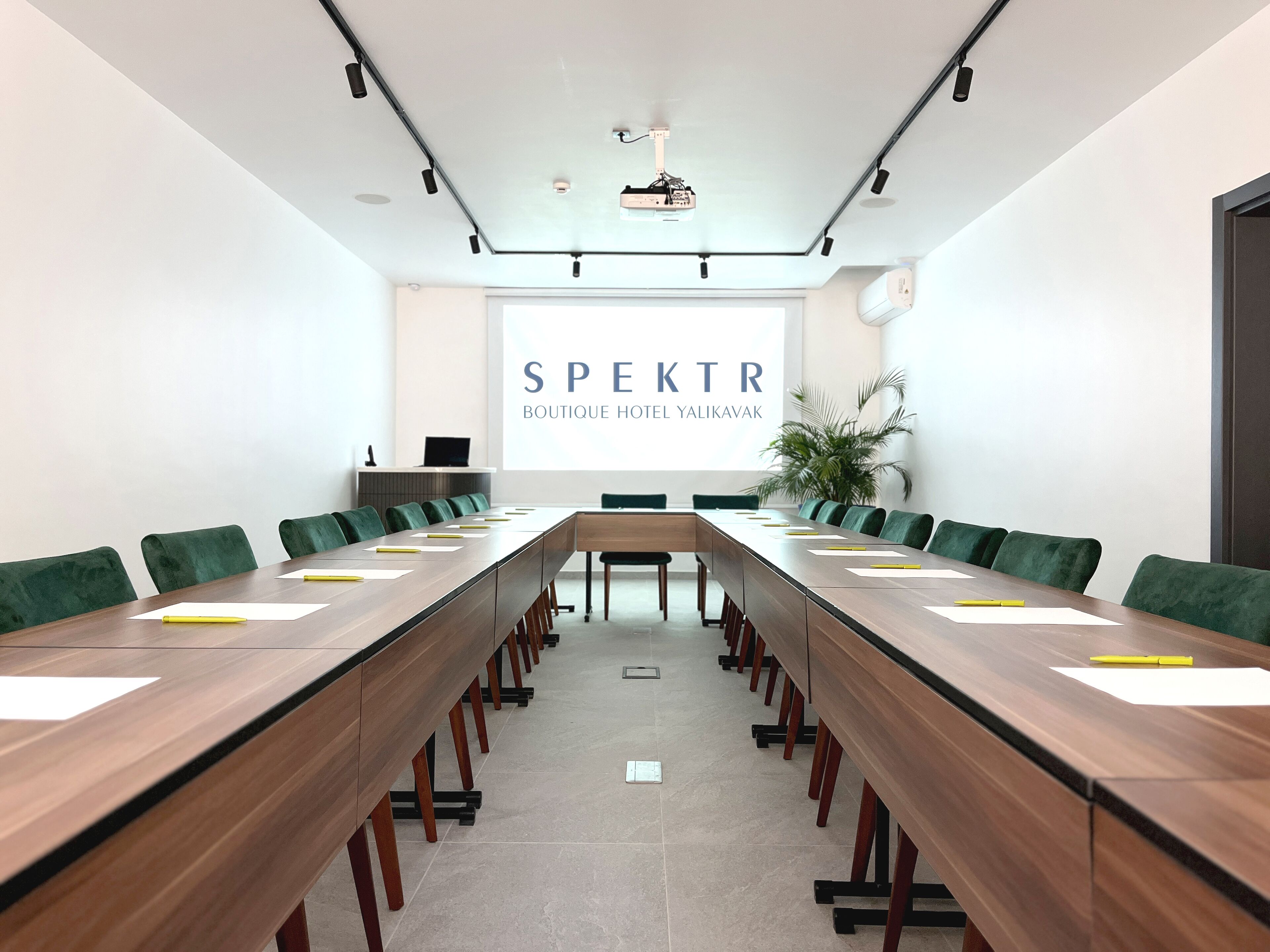 A conference room with a long table, green chairs, a projector screen, and a plant.