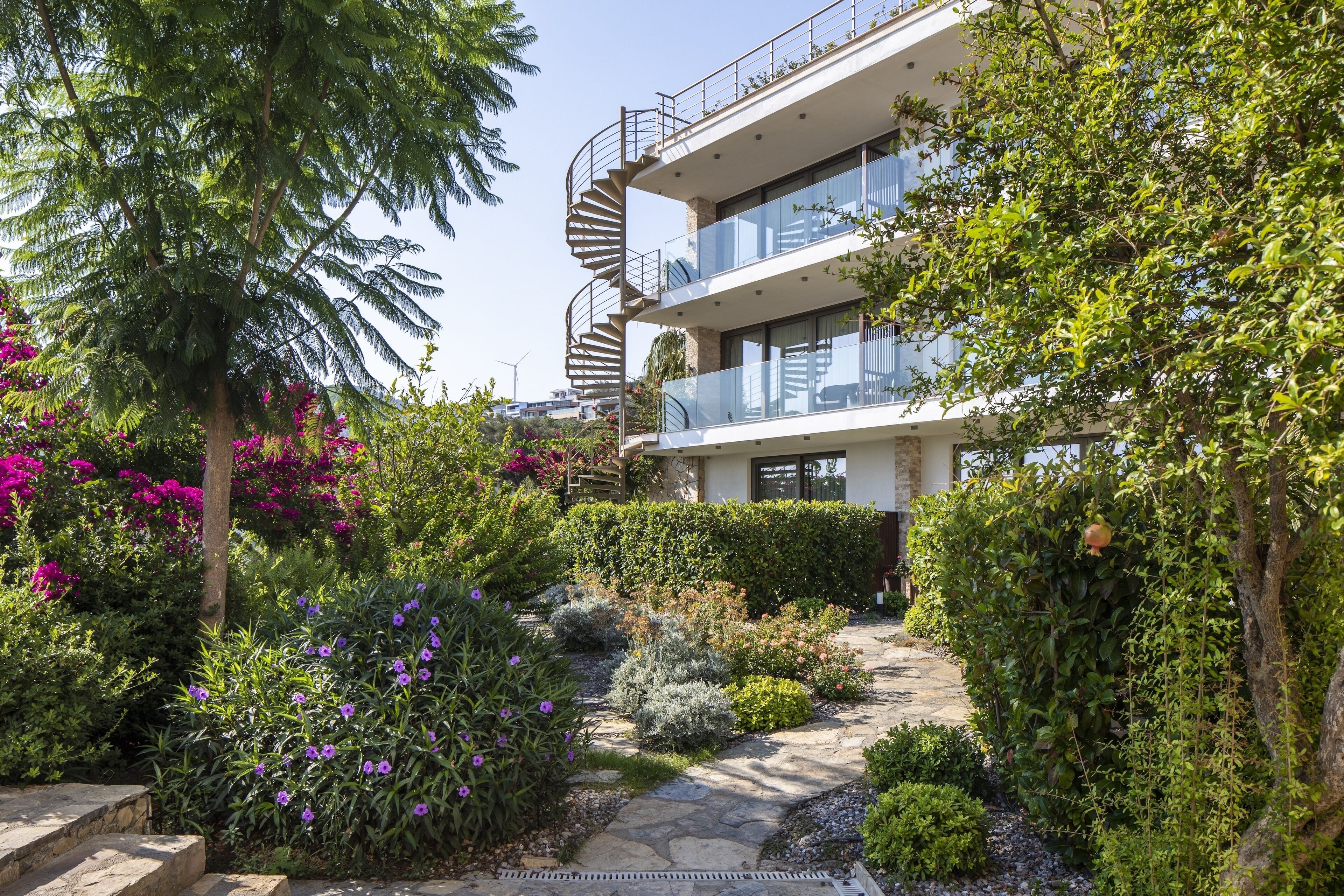 A modern multi-story building with a spiral staircase, surrounded by lush greenery and flowering plants.