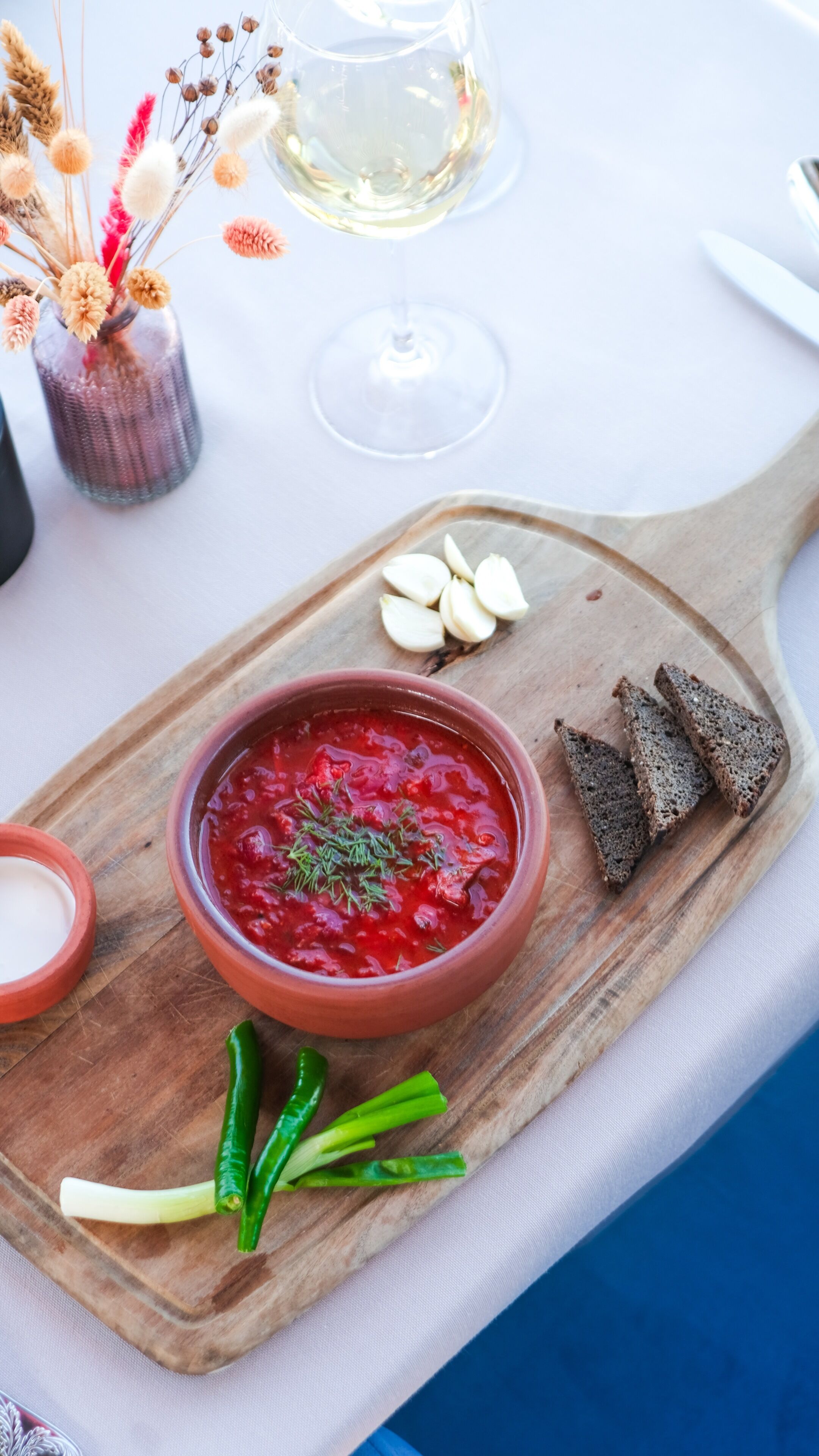 A wooden serving board with a bowl of red soup garnished with herbs, accompanied by slices of dark bread and garlic cloves.