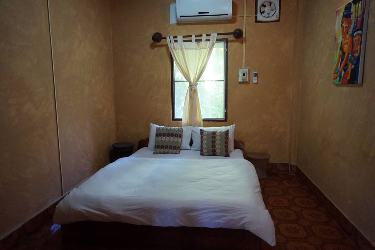 A small, cozy bedroom with a bed, two pillows, a window with a curtain, an air conditioner, and a wall-mounted fan.