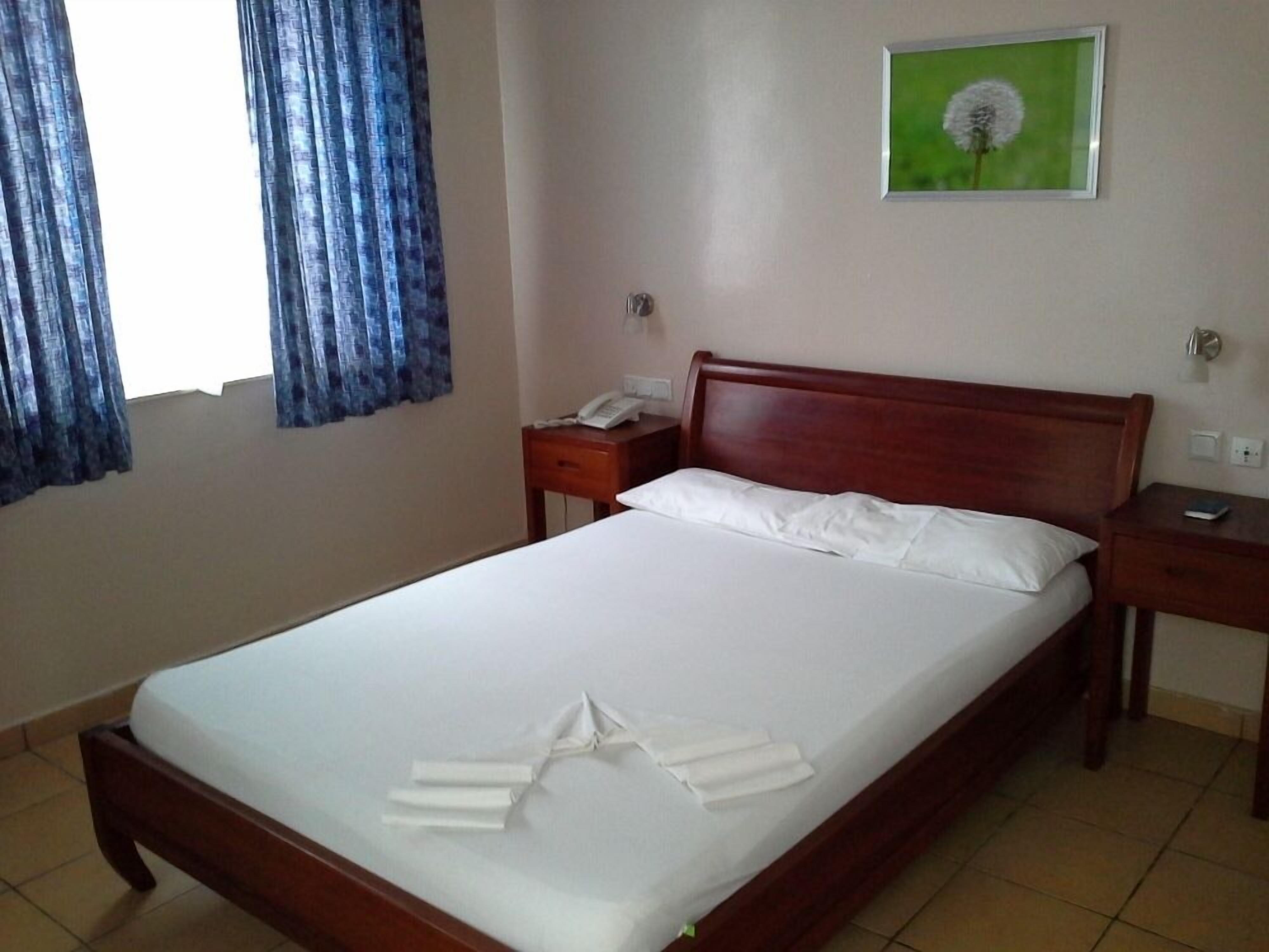 Un lit bien fait, avec du linge de lit blanc et une serviette joliment disposée, une tête de lit en bois, une table de chevet avec un téléphone, une petite table de nuit et un tableau encadré de vert accroché au mur.