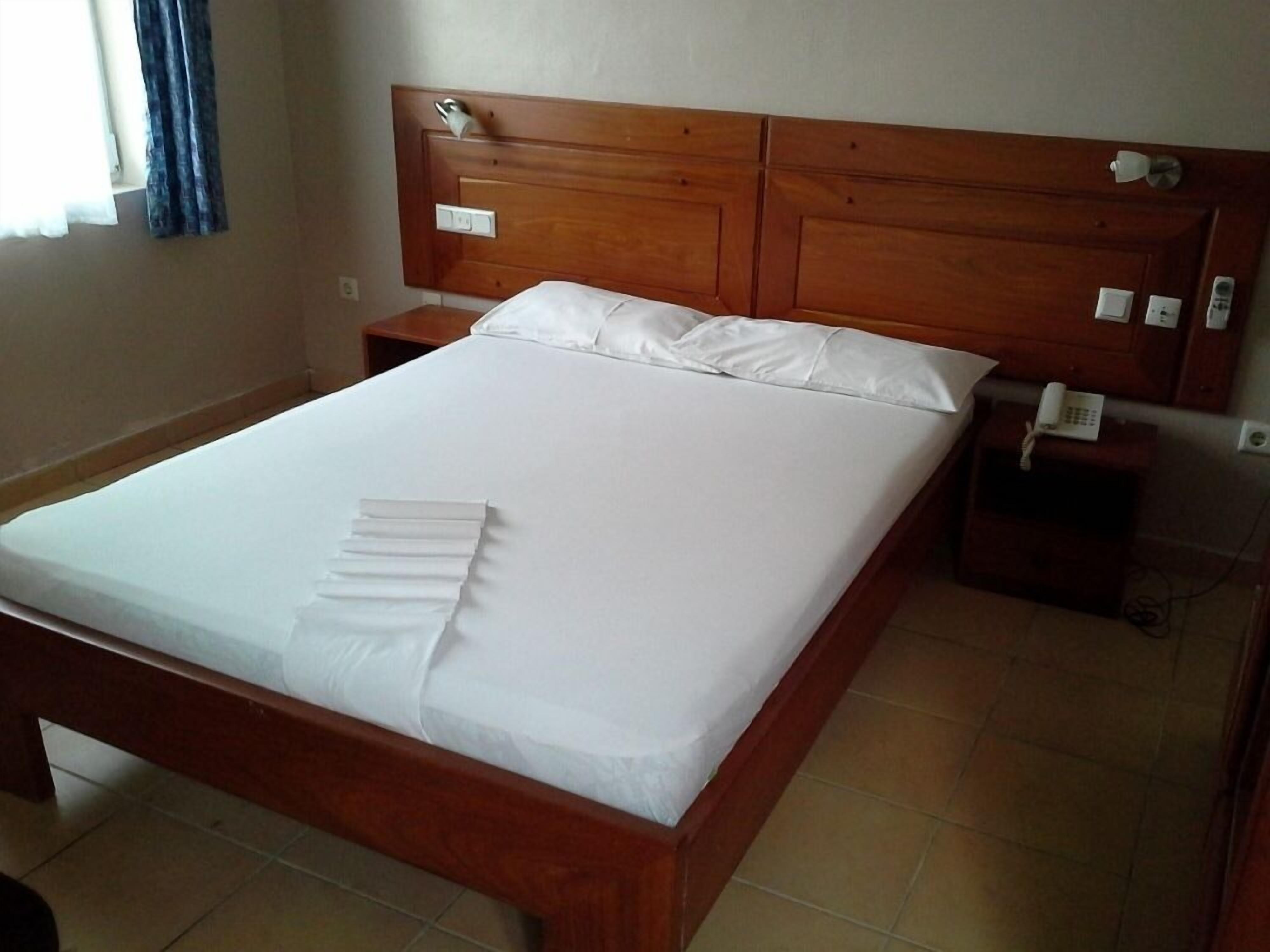 A standard hotel room with a wooden headboard, a neatly made bed with white linens, a bedside table with a phone, and a small nightstand.