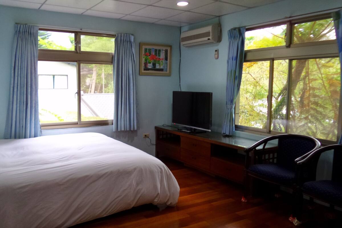 A hotel room with a bed, a desk with a television, and large windows with curtains.