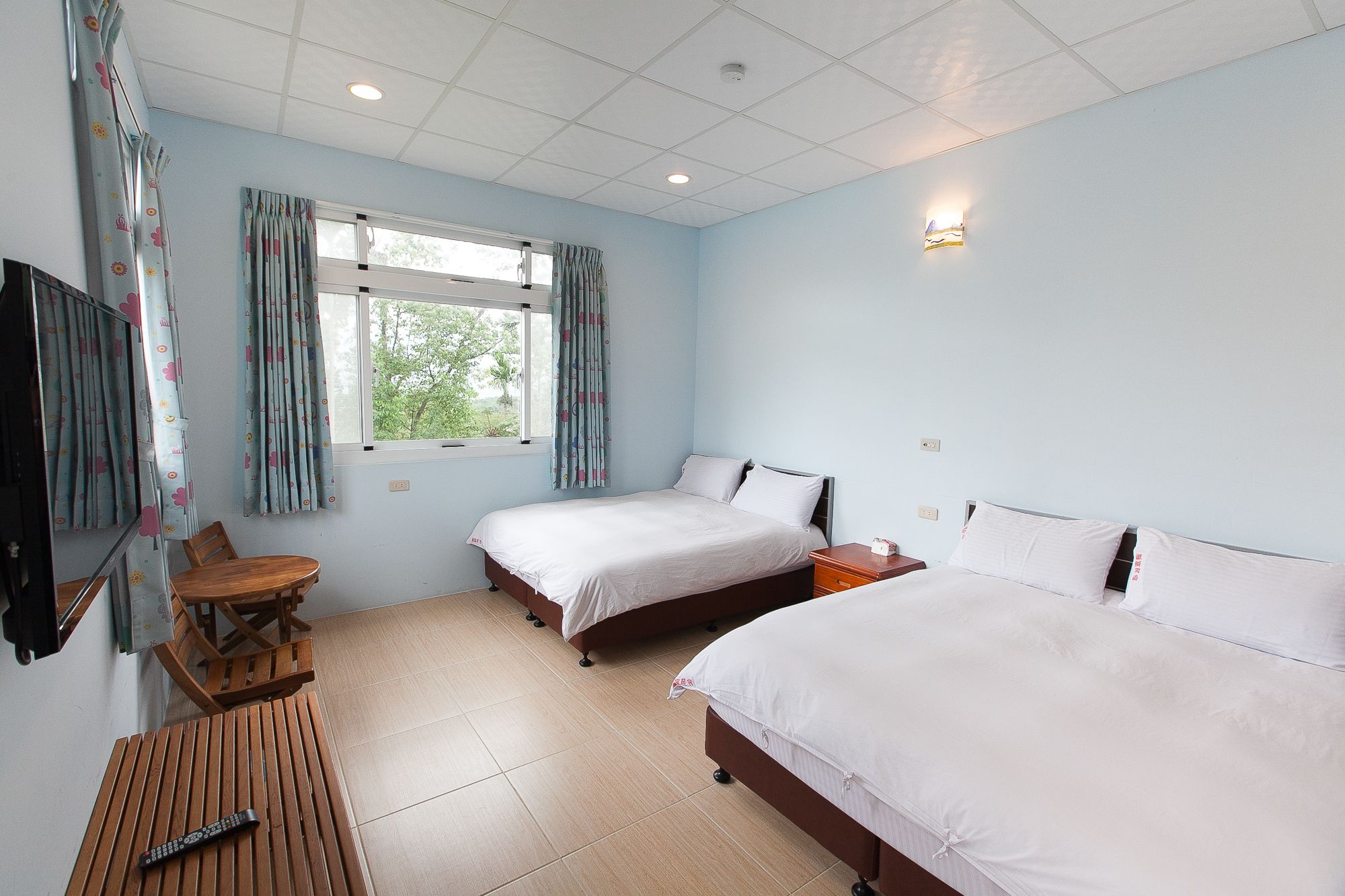 A bedroom with two beds, a wooden table, a TV, and a window with curtains.
