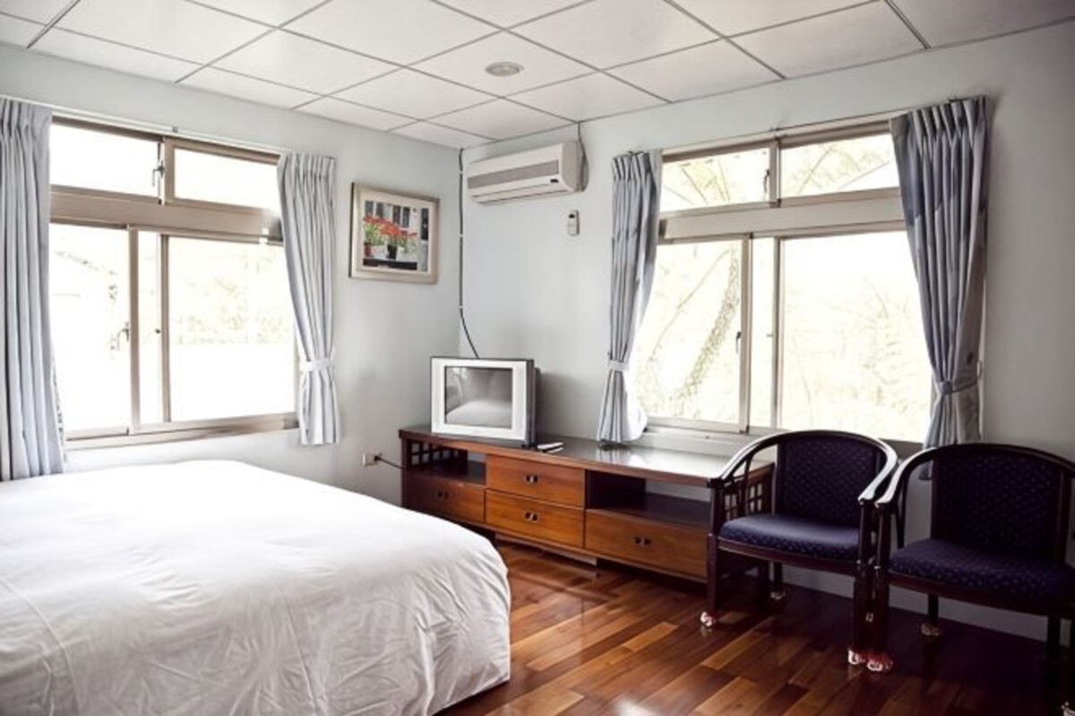 A bedroom with a bed, a desk with a television, two chairs, and large windows with curtains.