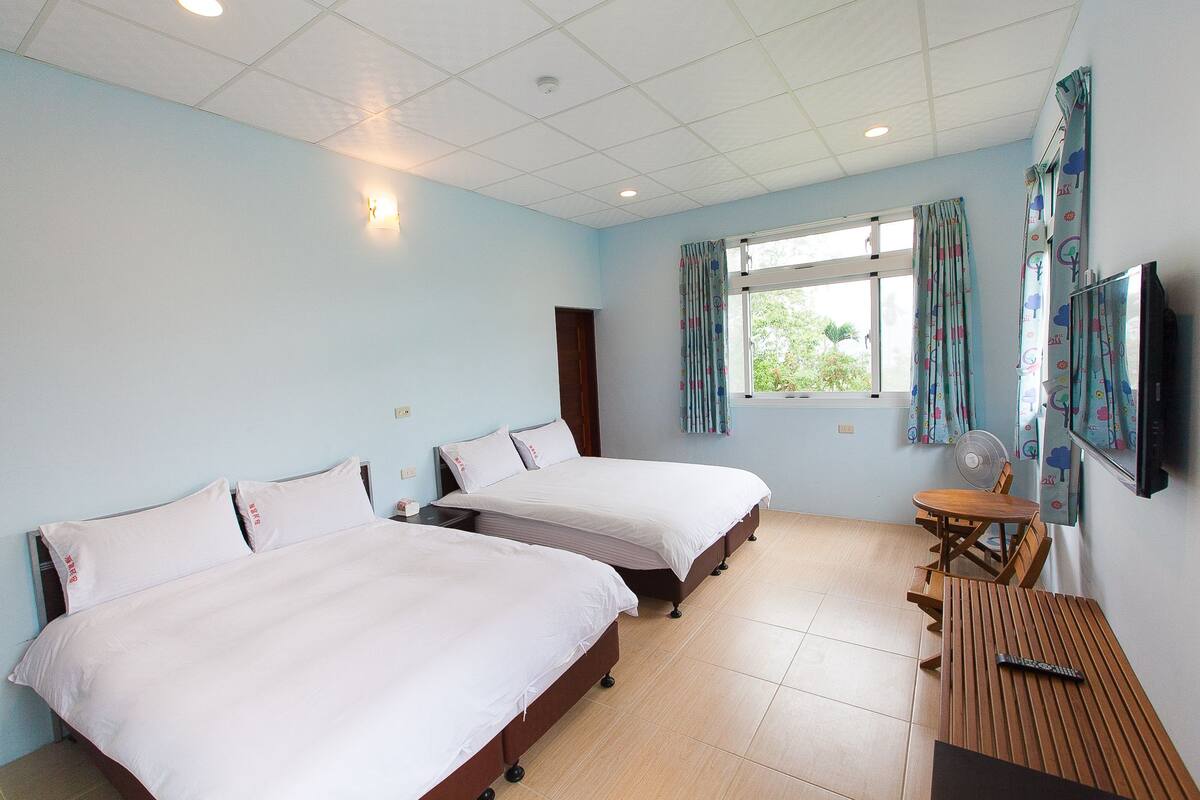A hotel room with two beds, a wooden table, a TV, and a window with curtains.