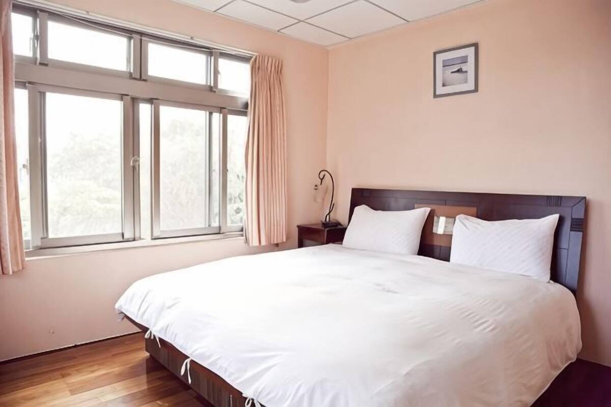 A bedroom with a large bed, a nightstand, a window with curtains, and a framed picture on the wall.