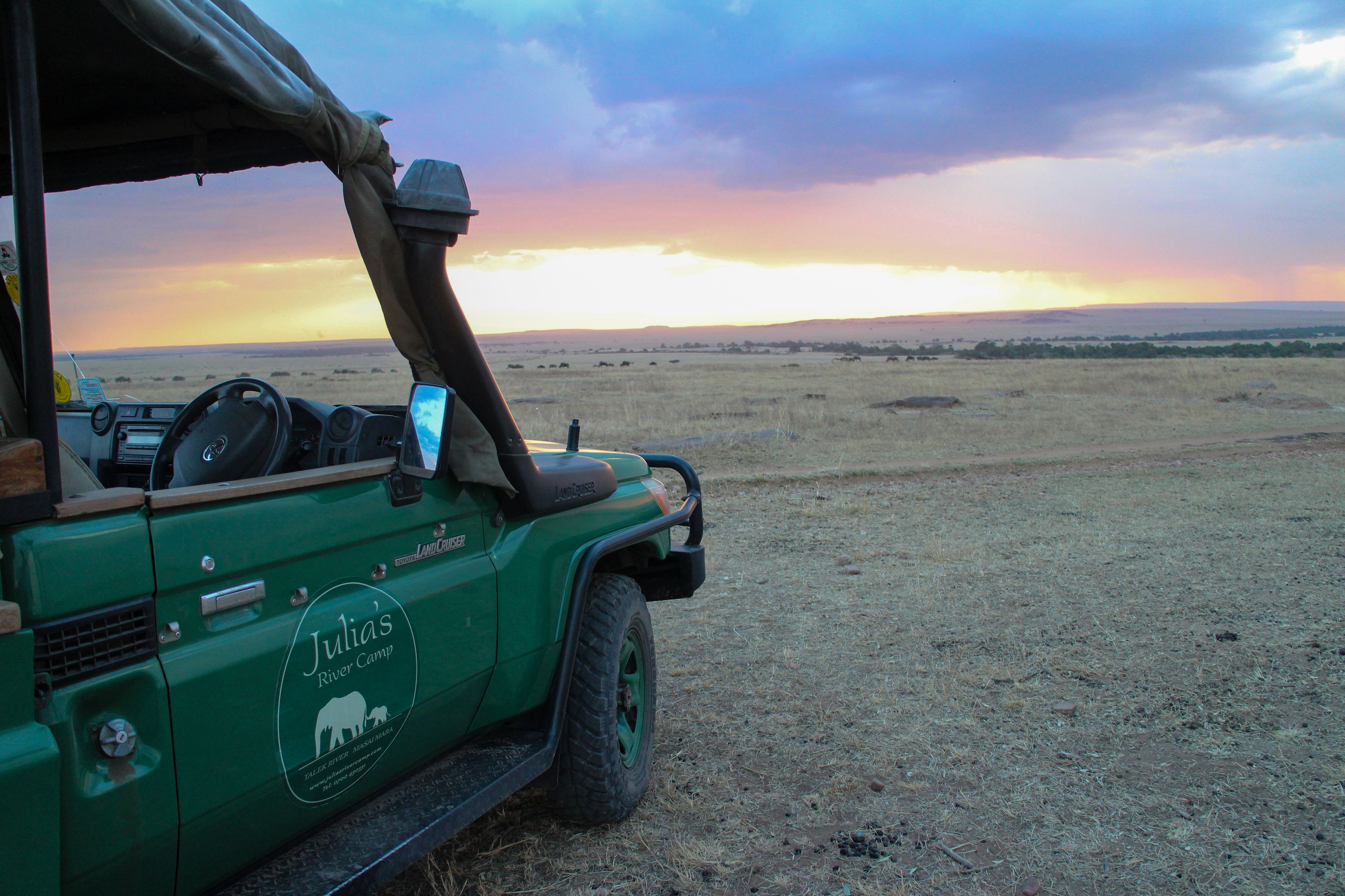 A green safari vehicle with "Julia's River Camp" branding parked in a dry, open field with a sunset in the background.