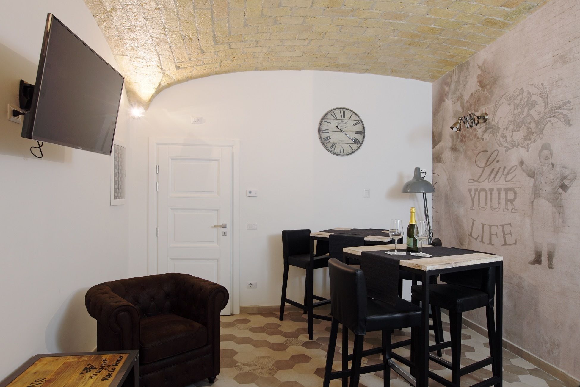 A cozy room with a flat-screen TV mounted on the wall, a round wall clock, and a table with chairs.