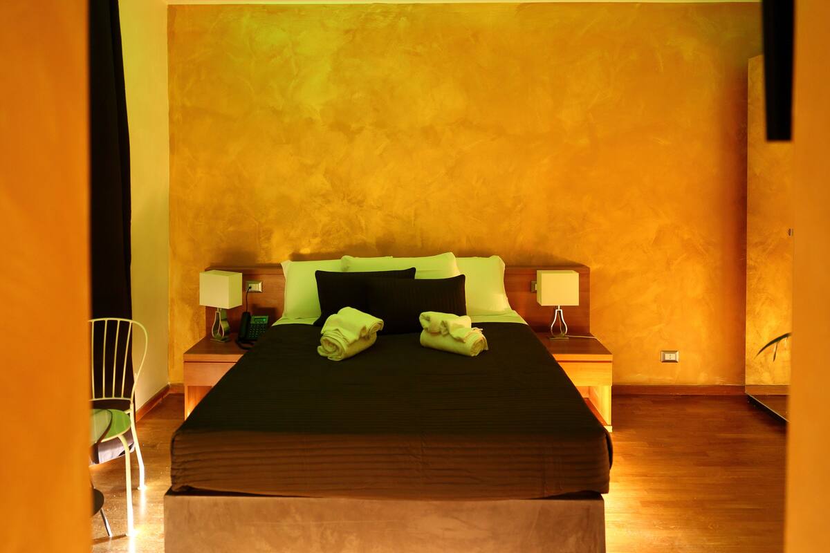 A hotel room with a large bed, two bedside tables with lamps, a chair, and a wooden floor.