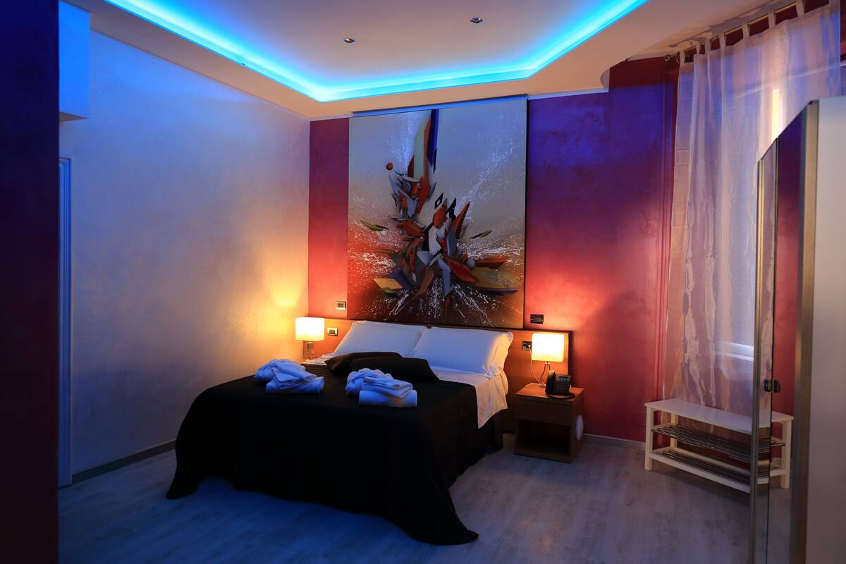 A modern hotel room with a large bed, two bedside lamps, a painting on the wall, and a small bedside table.