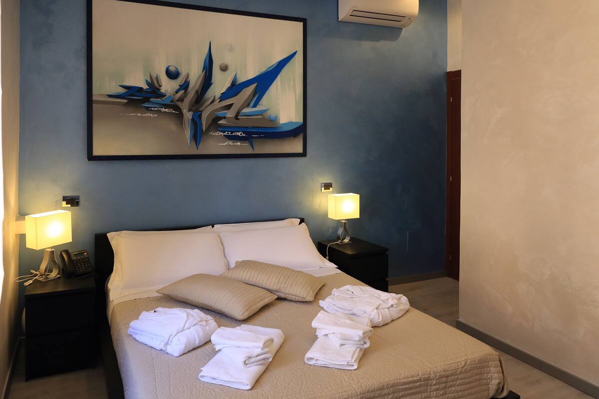 A neatly made bed with pillows and folded towels, a painting on the wall, and two bedside lamps.