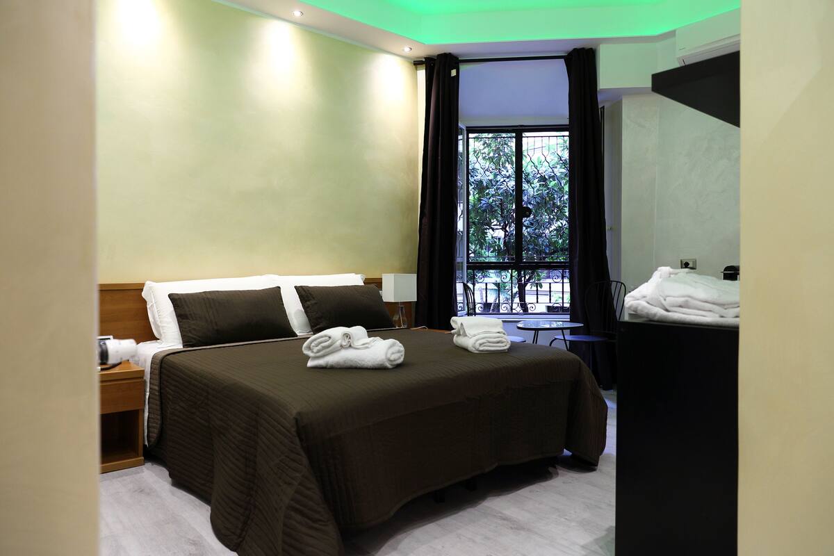 A hotel room with a large bed, two pillows, a nightstand, a desk, and a window with a view of greenery.