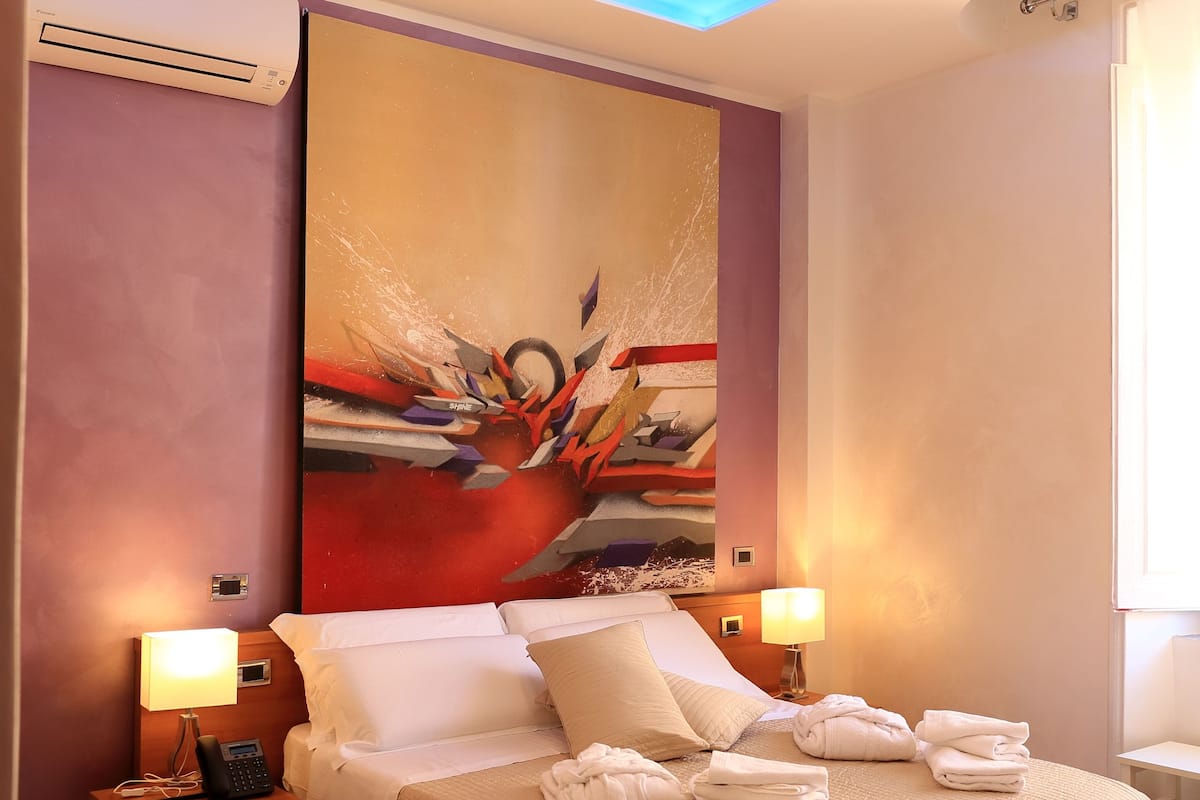 A modern hotel room with a large bed, two bedside tables with lamps, a ceiling with blue LED lighting, and a large abstract painting on the wall.