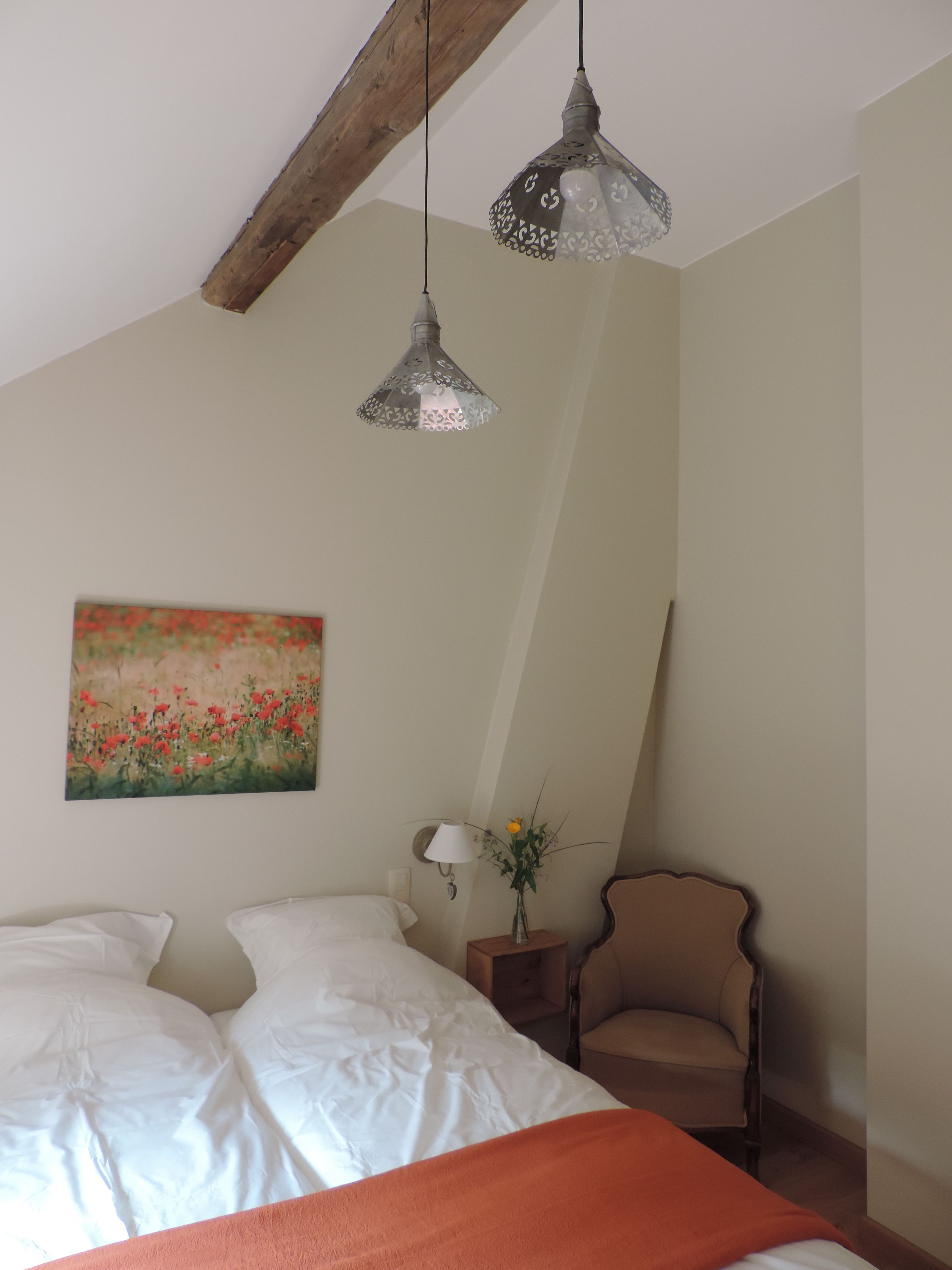 A bedroom with a bed, two hanging lamps, a chair, a nightstand, a vase with flowers, and a painting on the wall.