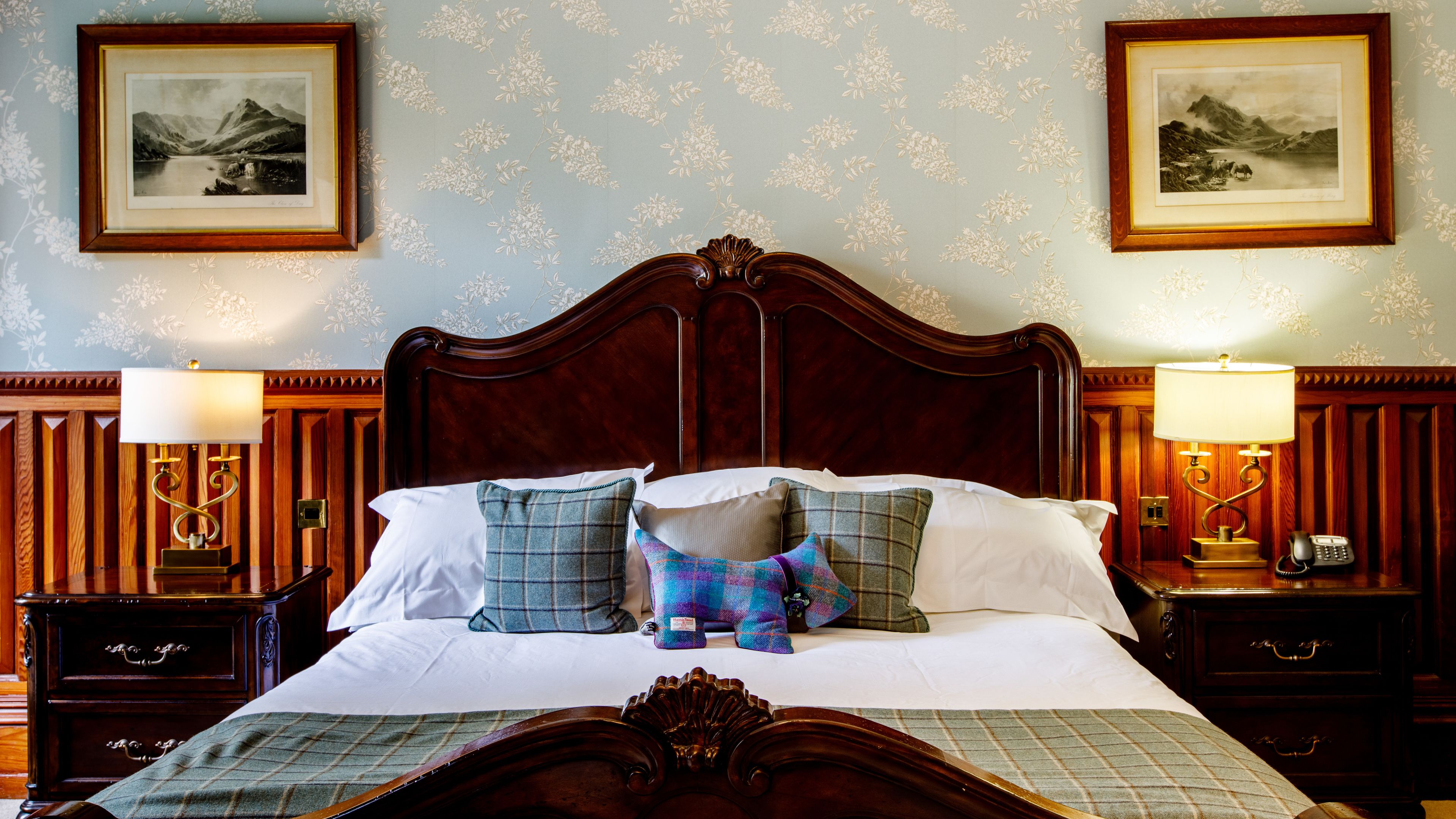 A neatly made bed with a wooden headboard, flanked by two bedside tables with lamps and a telephone. Two framed landscape paintings adorn the walls above the bed.