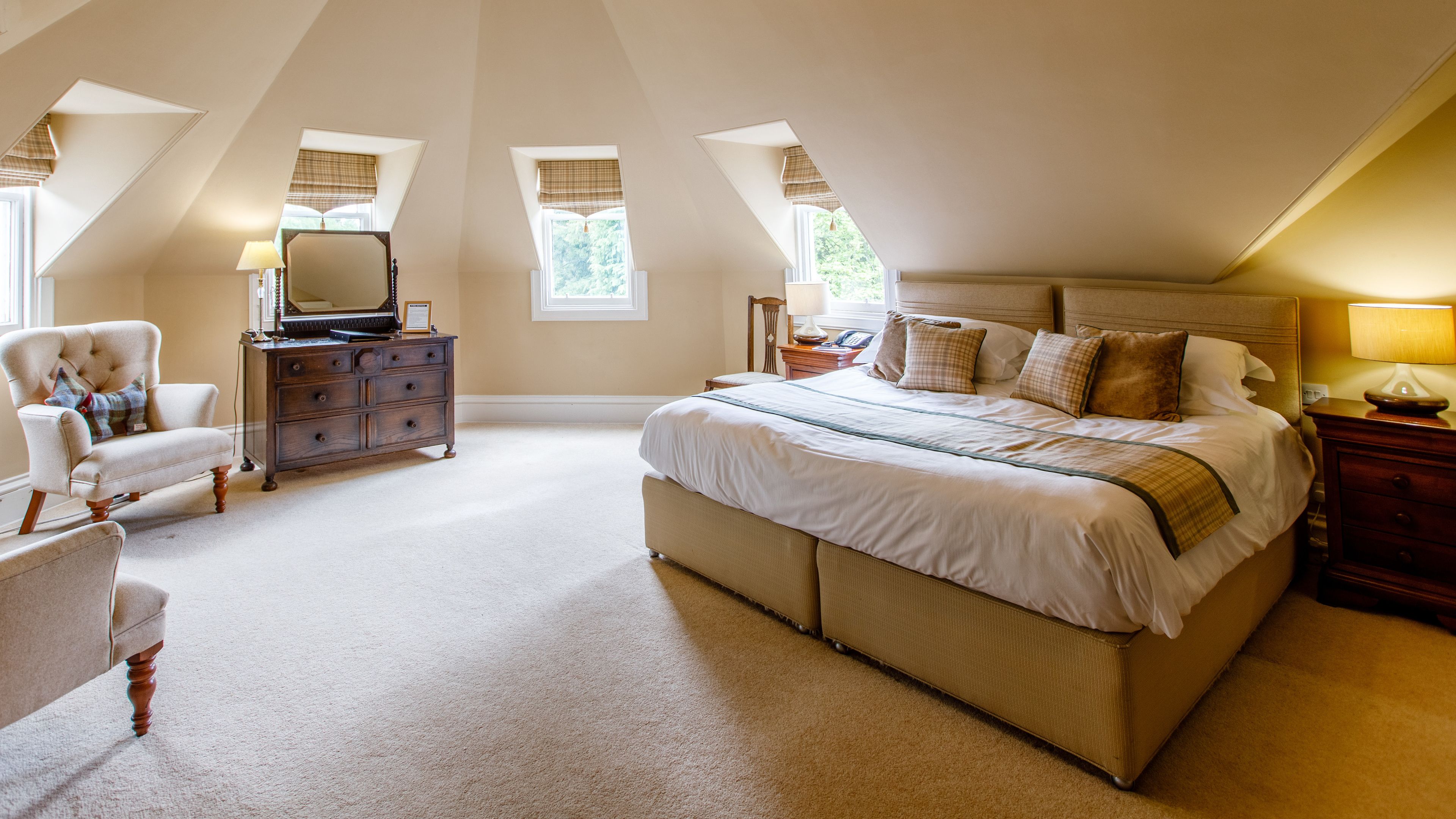 Une chambre spacieuse avec un grand lit, une commode, une chaise et un bureau. Elle est dotée de deux fenêtres avec des rideaux et possède un plafond en pente.