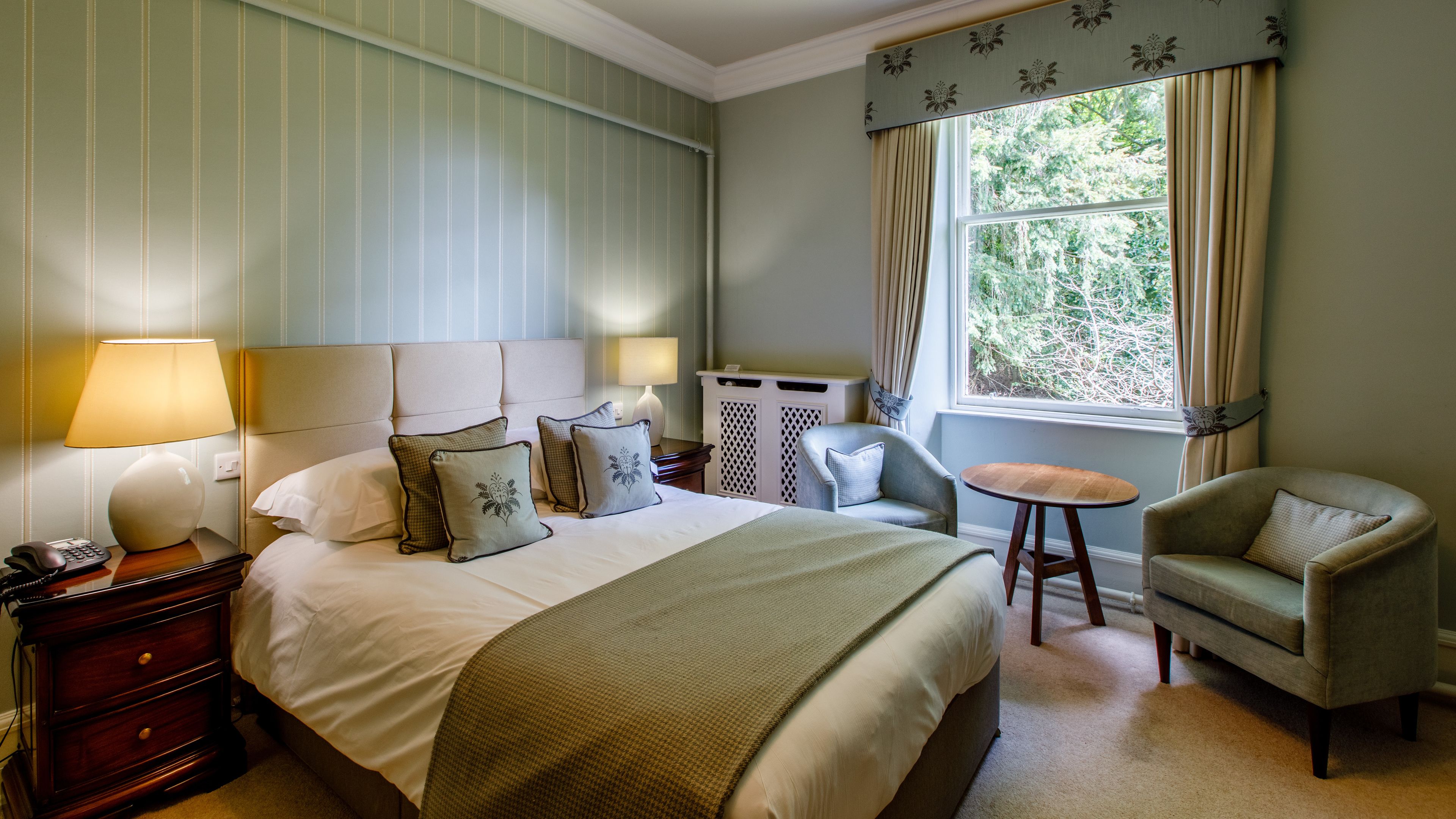 A hotel room with a bed, a chair, a small table, a lamp, and a window with curtains.
