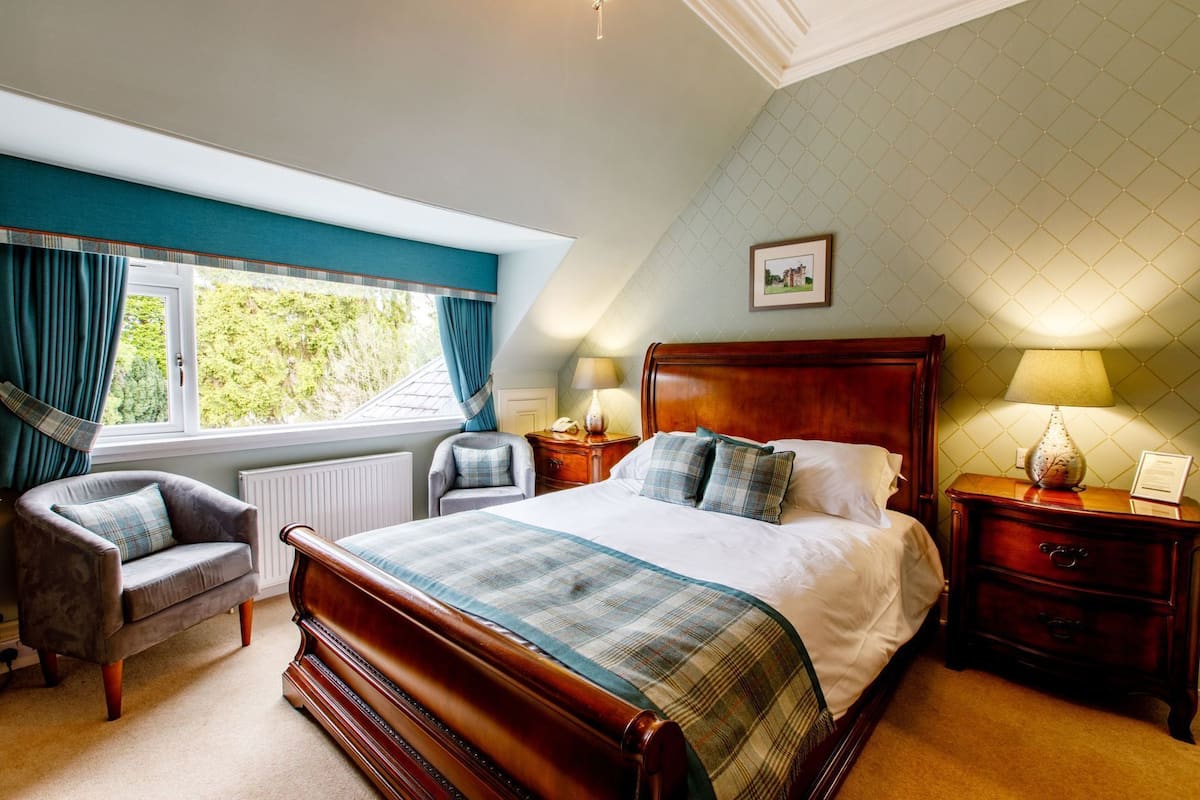 A bedroom with a large bed, a chair, a nightstand, a lamp, and a window with curtains.